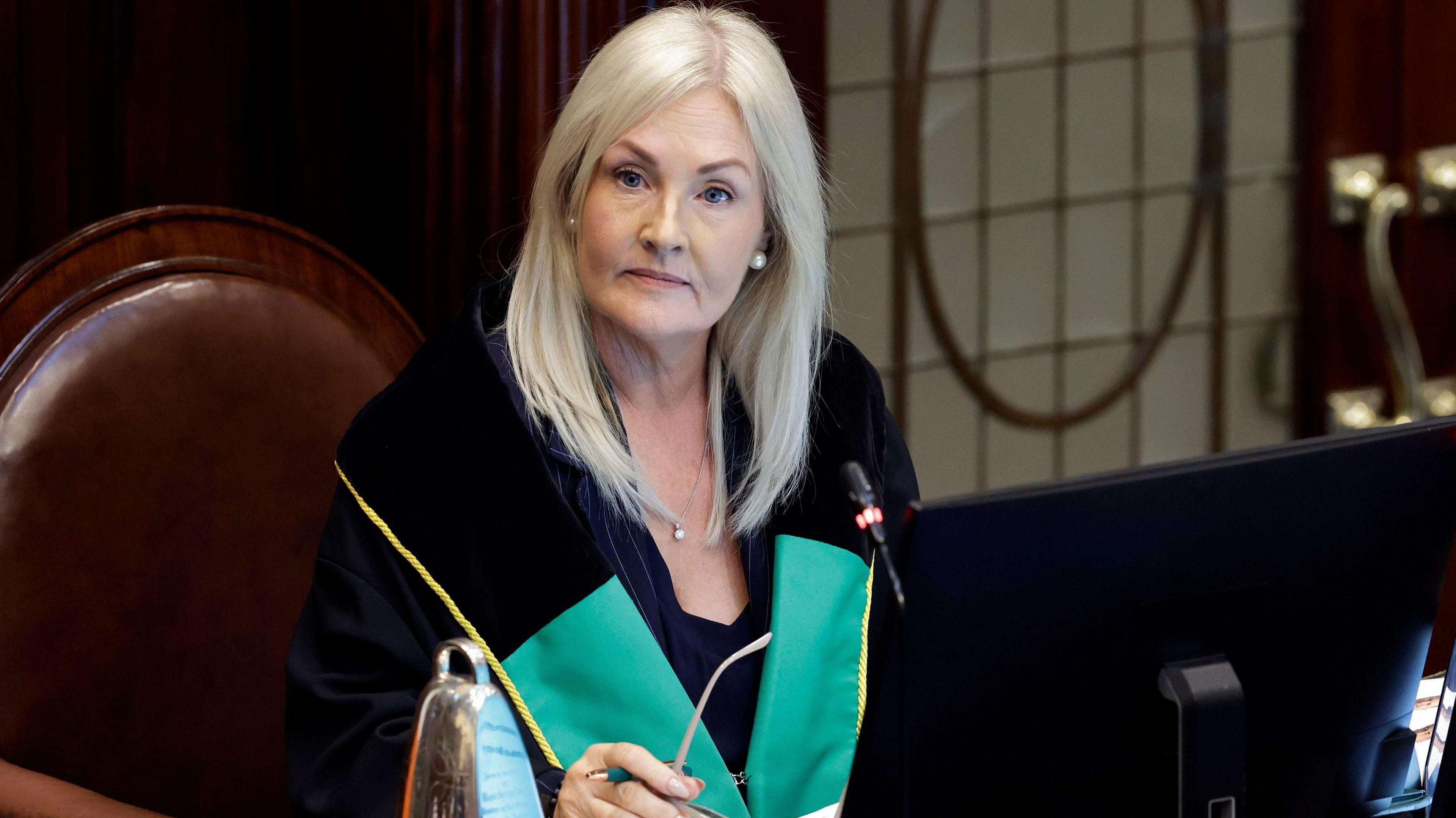 Verona Murphy has long light blonde hair and is wearing a green and black robe while acting as speaker in the Irish parliament. She is sitting at a desk on a brown leather chair with a pen in her hand.