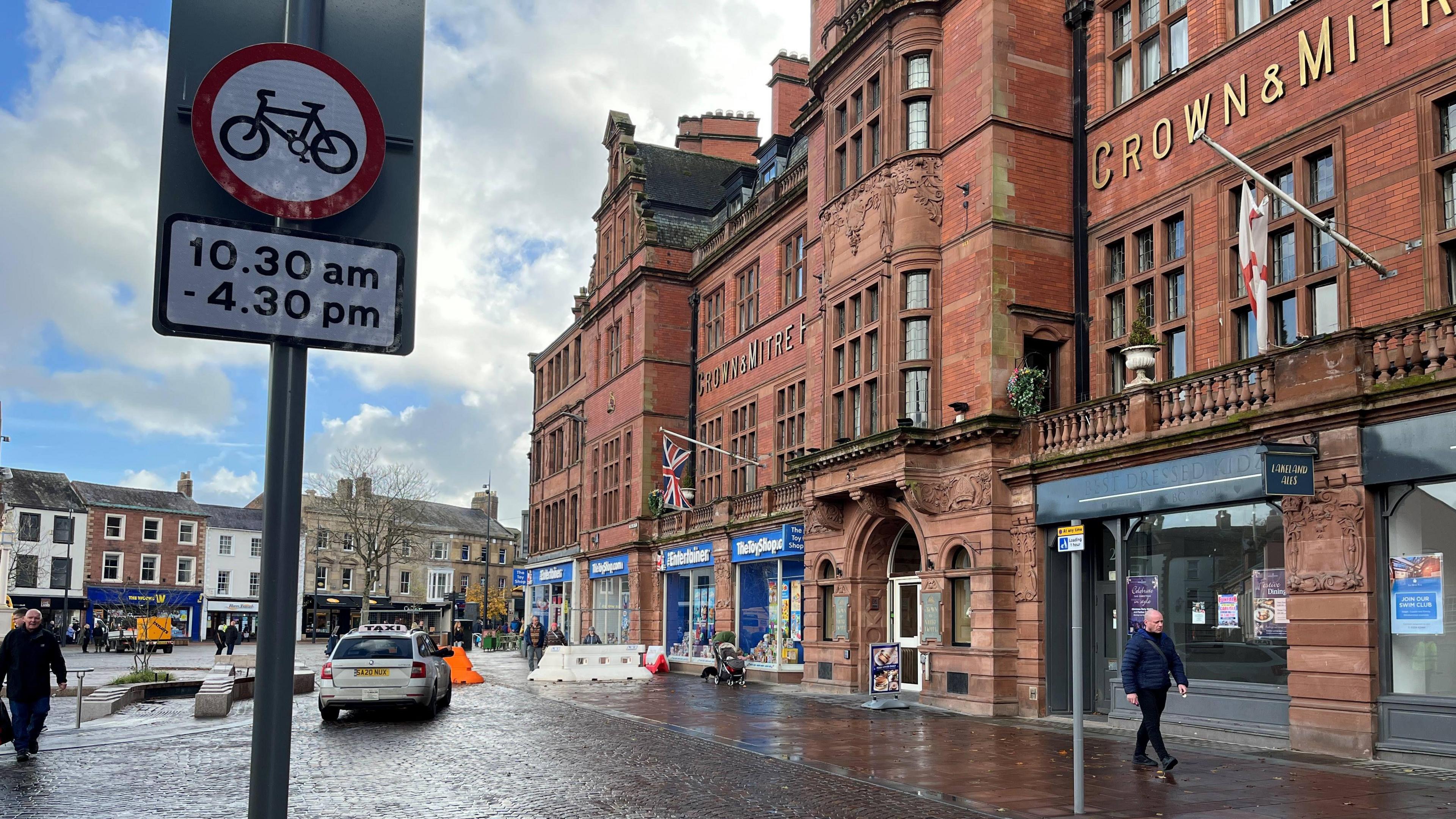 The entrance to the pedestrian area by the Crown and Mitre in Carlisle, which is a large and beautiful redbrick building with leaded windows, a first floor balcony running most of its length, ornate stone work and many chimneys. The ground floor is given over to shop fronts. A taxi is parked in front of an orange bollard, behind a sign banning cyclists from 10.30am to 4.30pm.