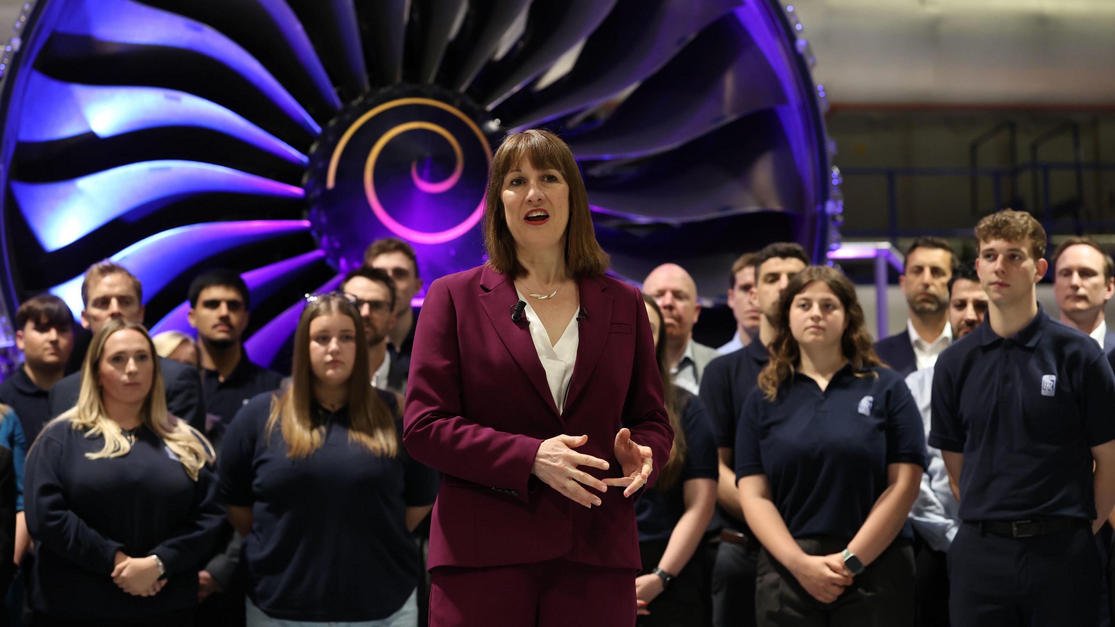 Rachel Reeves, in a burgundy suit, giving a speech. In the background there is a group of Rolls Royce staff, looking at and listening to the Chancellor. Behind them, a large, silver jet engine is lit up by purple lights.