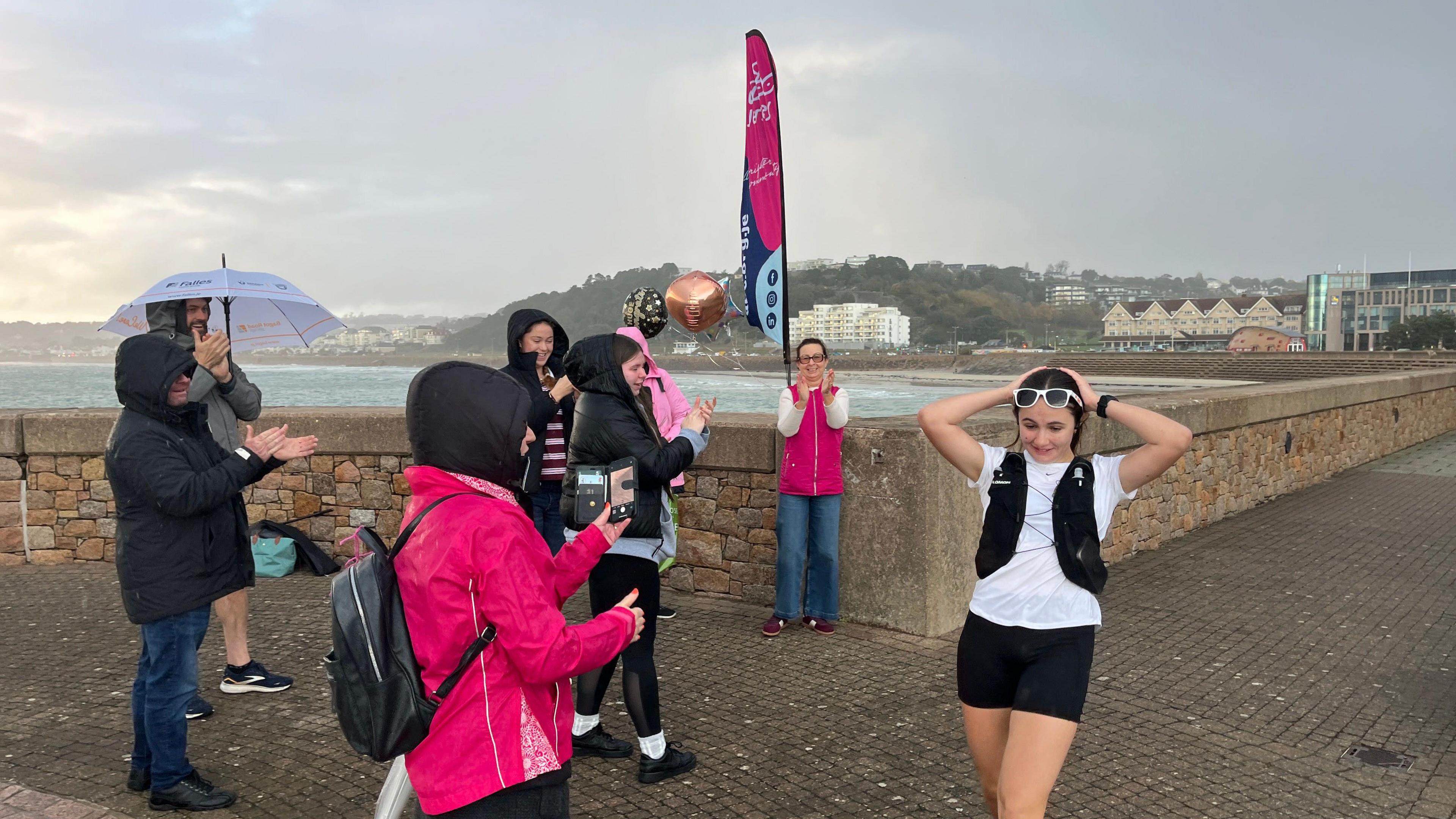 Emily is wearing a white shirt and black shorts with a hydration vest, she is in the foreground, hands on head. Several people in bright pink jackets and dark clothing stand nearby, some clapping and others taking photos. A pink event flag and a metallic balloon are visible, suggesting a celebratory moment. The background shows a stone seawall, water, and distant buildings under a gray, rainy sky.