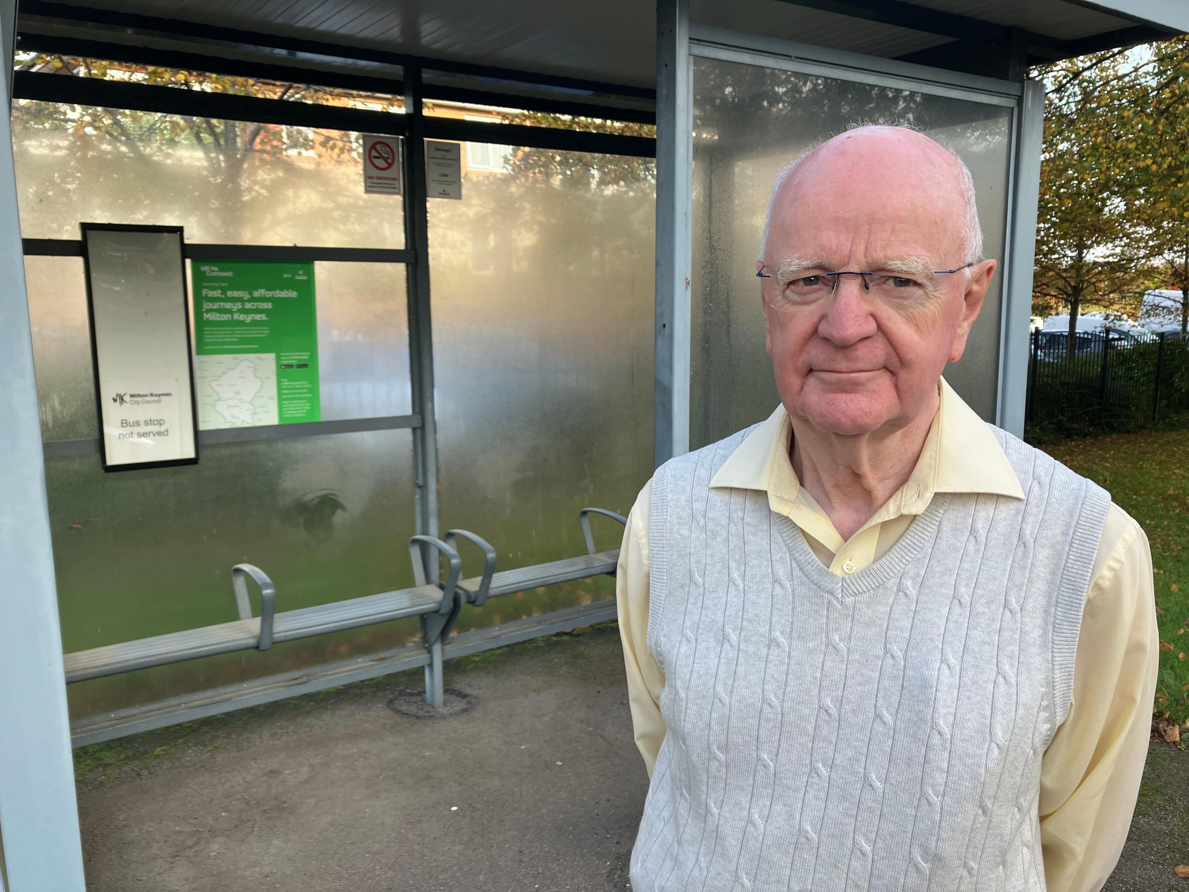 David Tunney who has white receding hair and glasses is standing in front of a bus stop with a 'bus stop not served' sign. He is wearing a light grey cricket-style, sleeveless jumper and a yelllow long sleeved shirt.