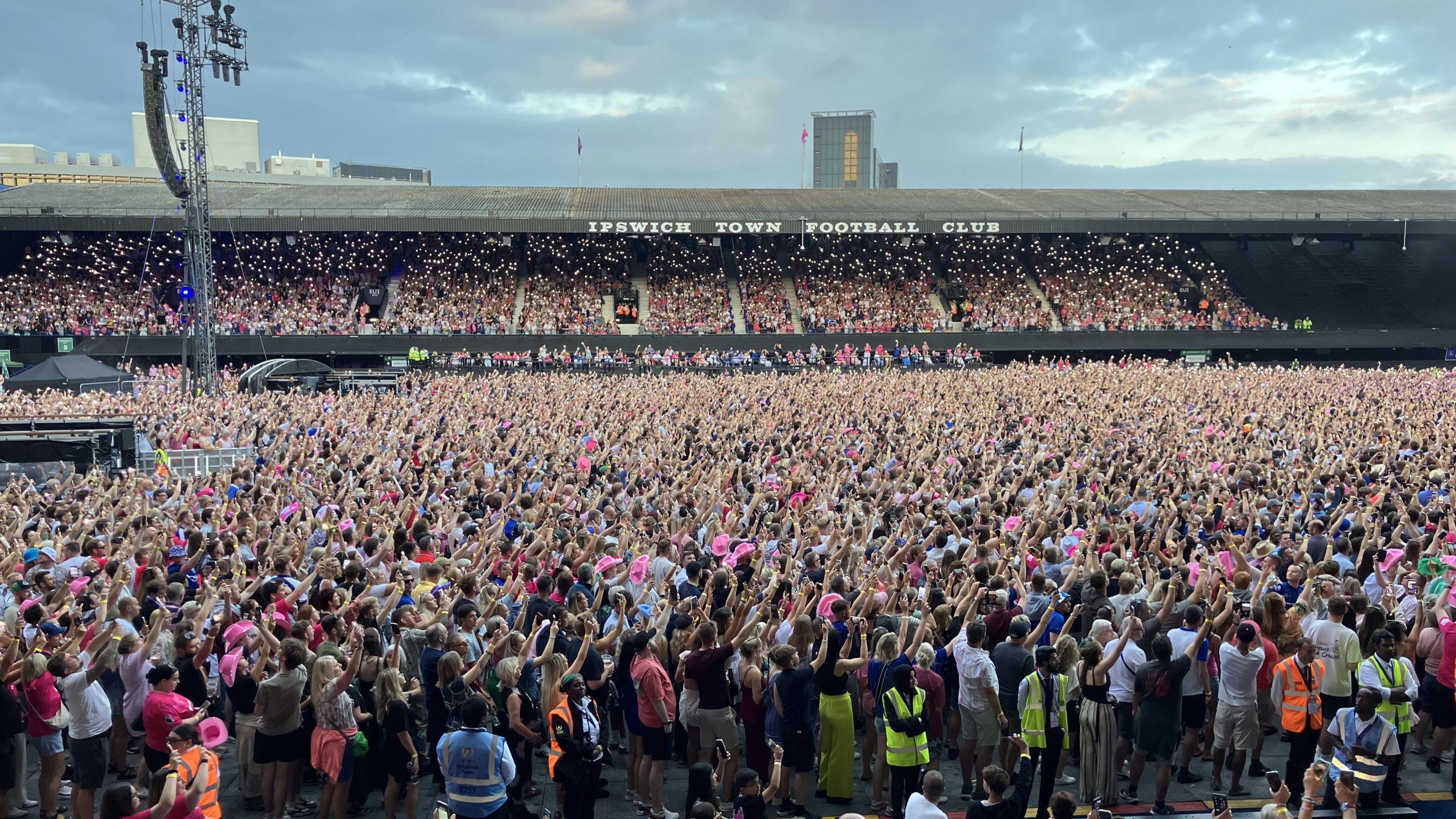 A view of the crowd in Portman Road stadium. The fans have their arms raised in the air as they look toward the stage.