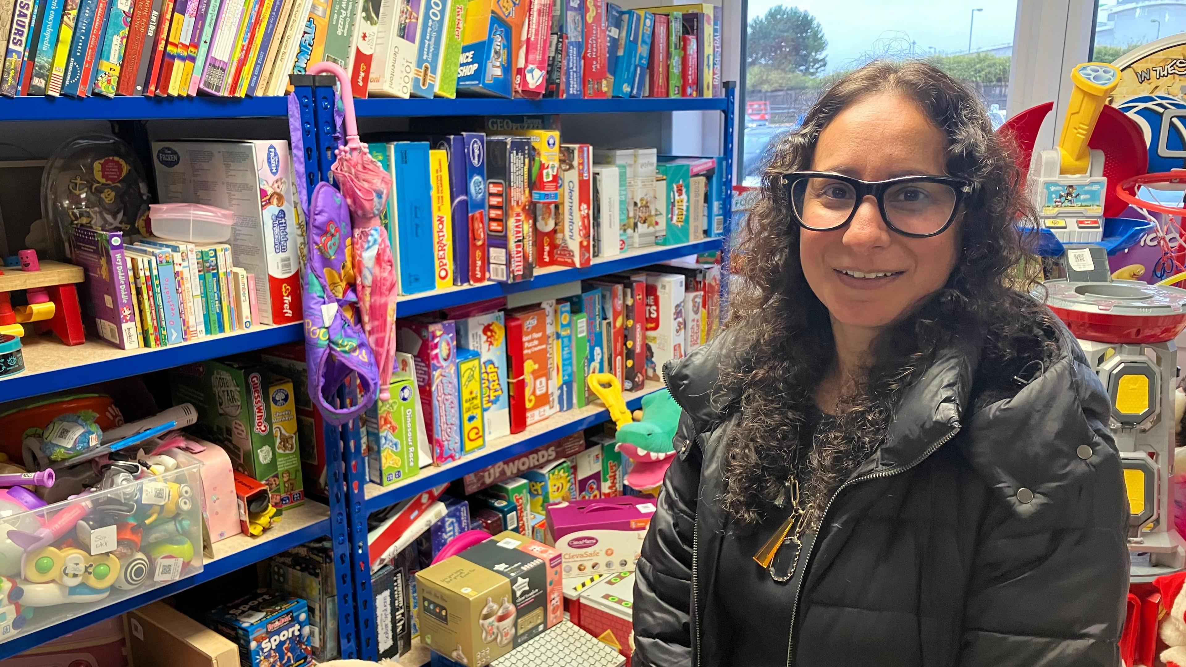 A woman in a black puffer jacket stands next to a four storey shelf with lots of children's books, board games and toys behind her.