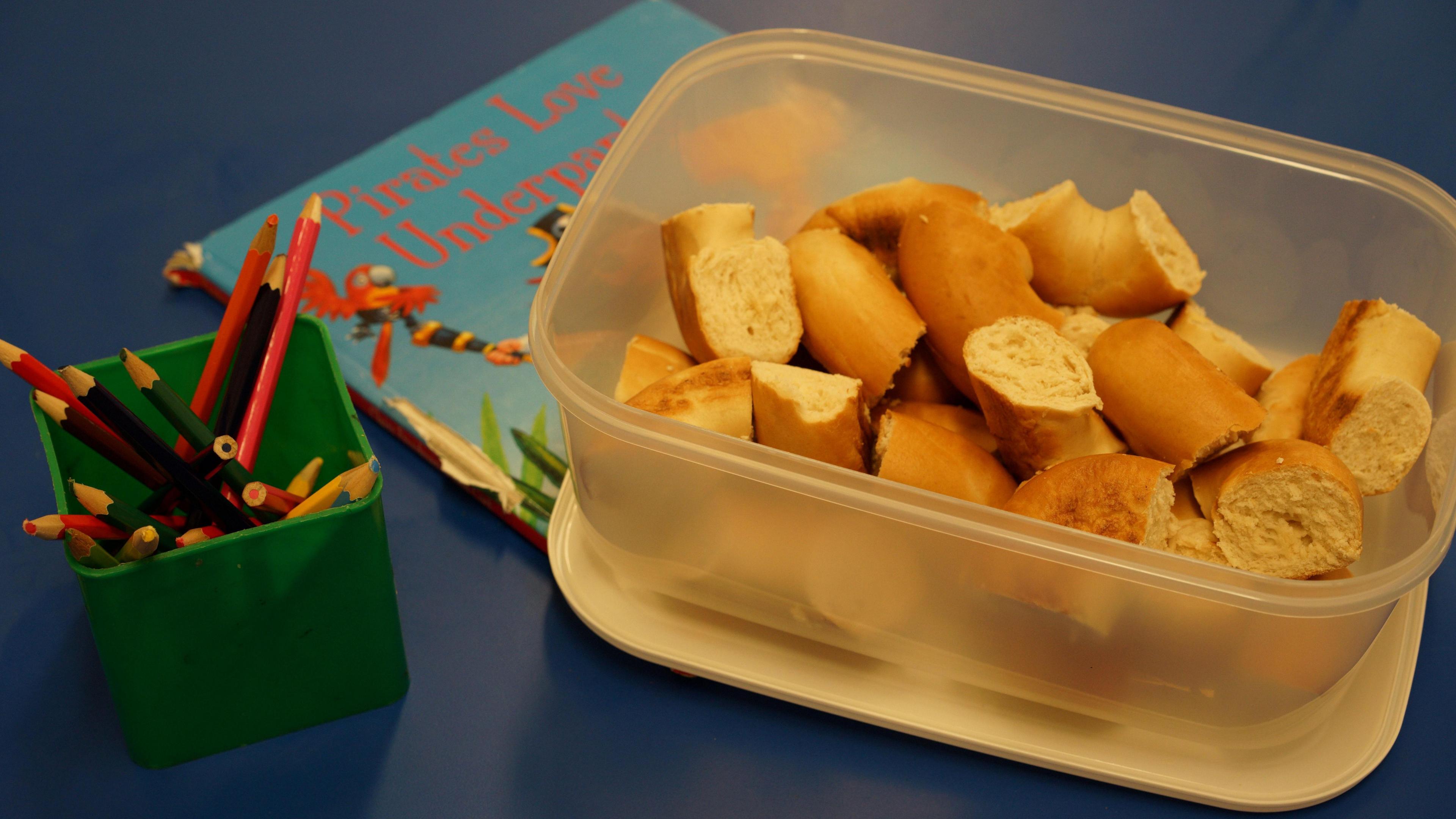 Pieces of bagel are in a plastic box on a blue table, there is also a green box holding some coloured pencil and a children's book.