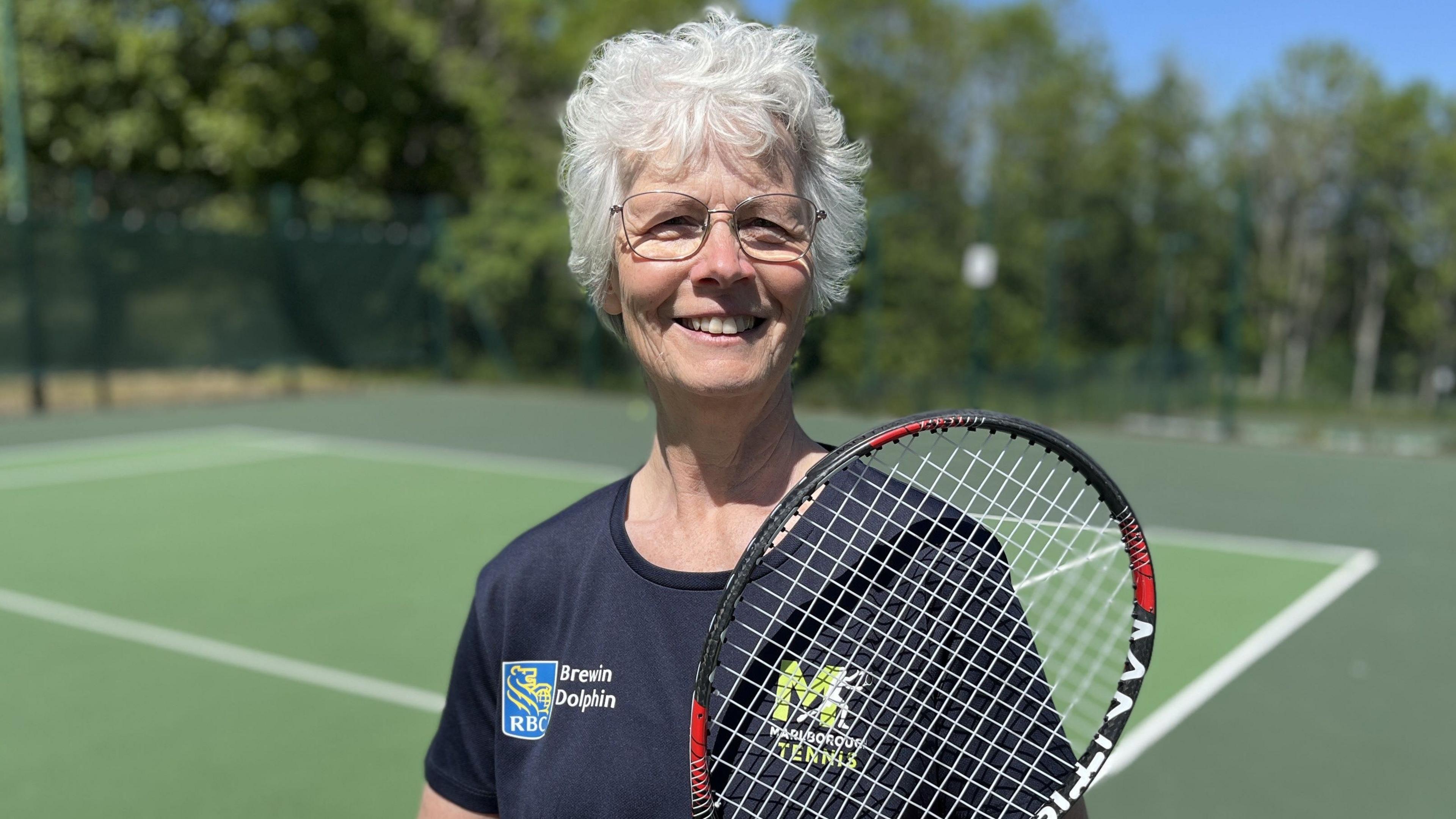Hilda Moore standing on court smiling wearing a Marlborough Tennis t-shirt holding her racket up to her shoulder