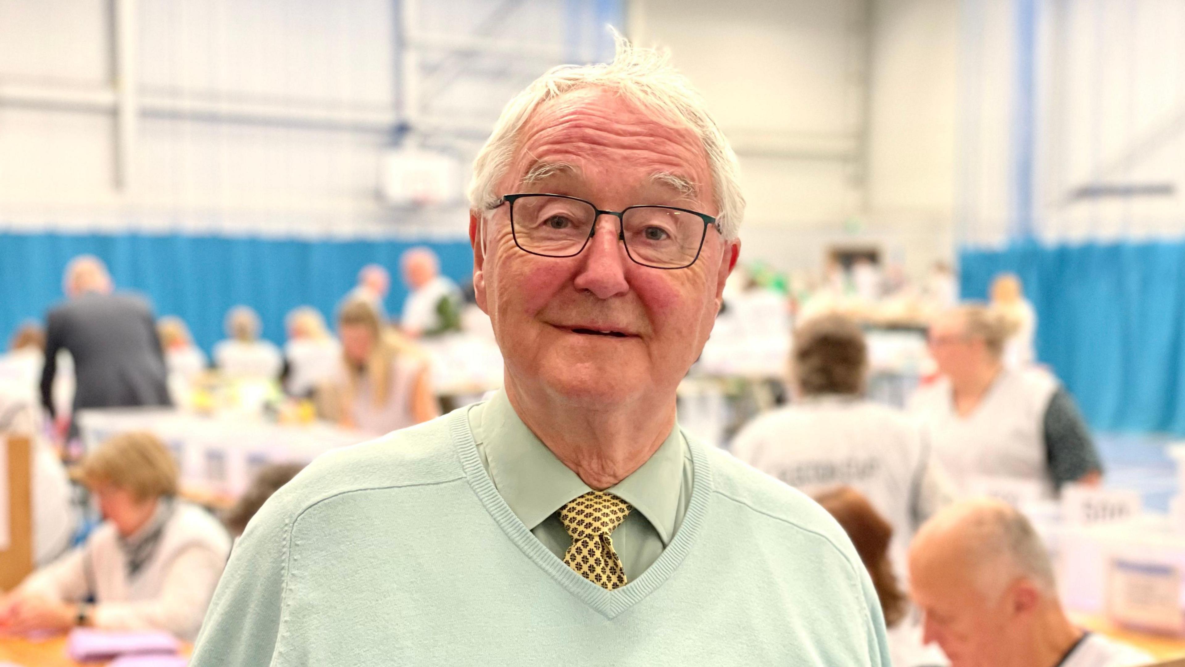 Councillor Roger Evans smiling, wearing glasses and a green pullover, at an election count.