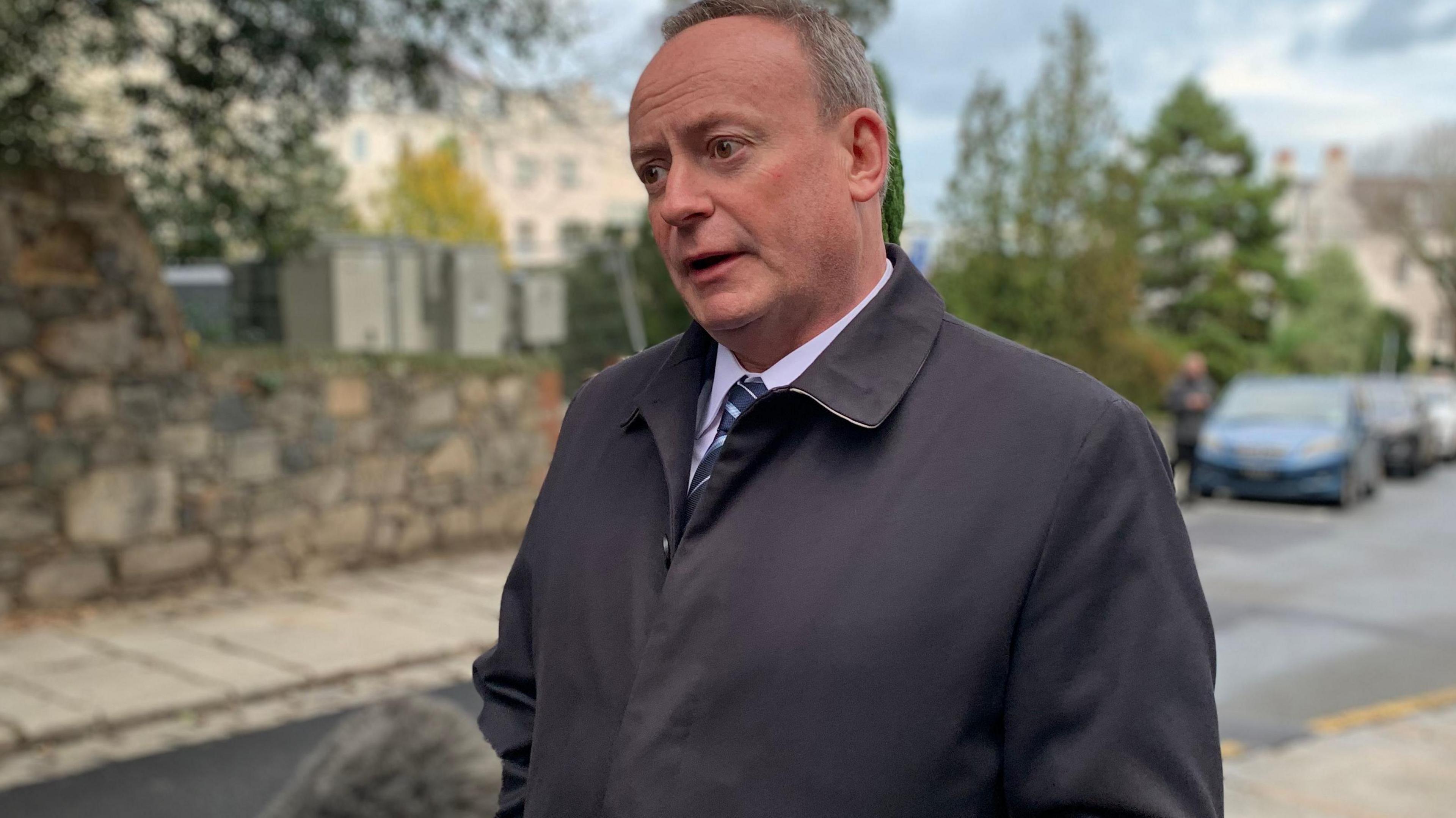 Deputy Lyndon Trott wearing a navy blue jacket with a light blue shirt and blue triped tie. He has grey, short hair. He is stood on a street with cars in the background.