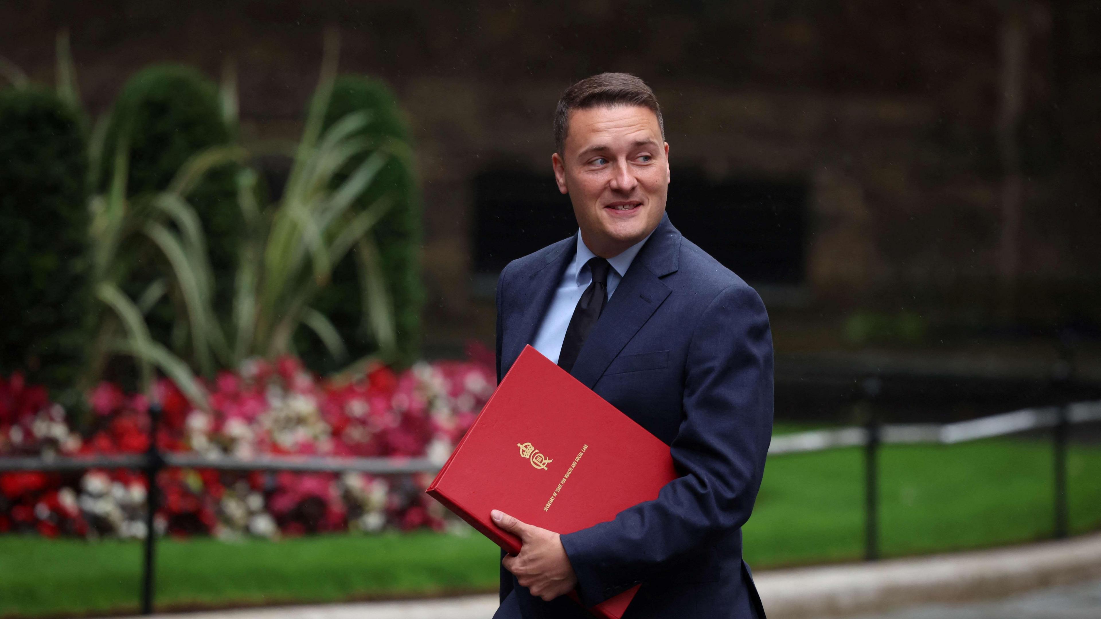 Wes Streeting holding a red folder is looking to his left, smiling, as he walks past some flowers and grass