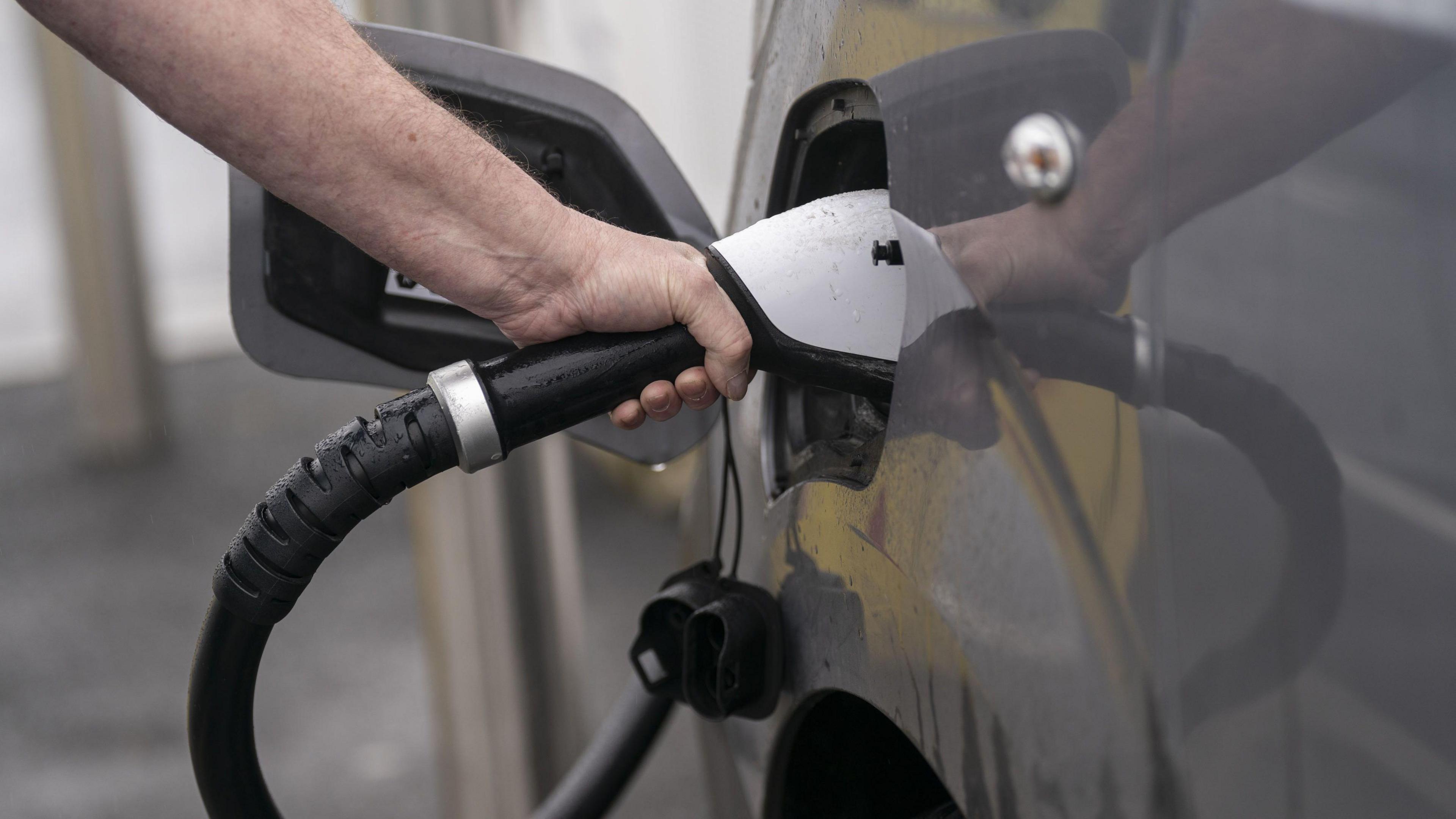 The image shows a hand inserting a white fuel pump nozzle into a grey car’s fuel filler door, with the fuel cap hanging below.