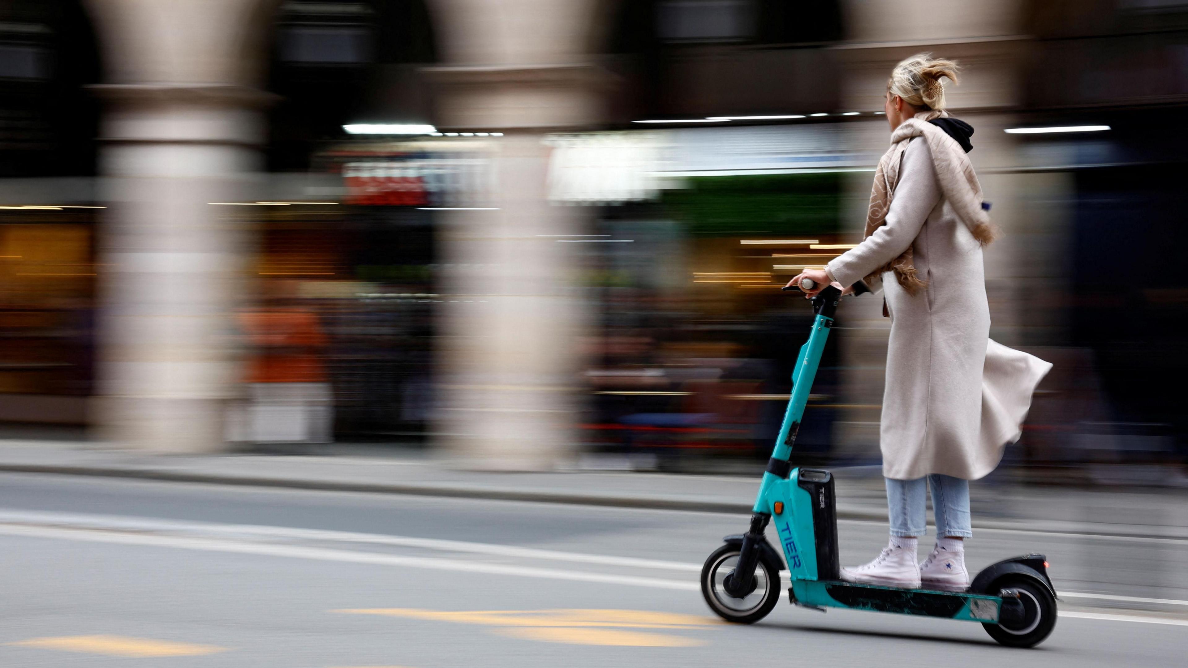 A woman riding on an e-scooter on the road. She is wearing a long cream-coloured coat and scarf, blue jeans and white high-top trainers. She has blonde hair clipped back with a clasp.