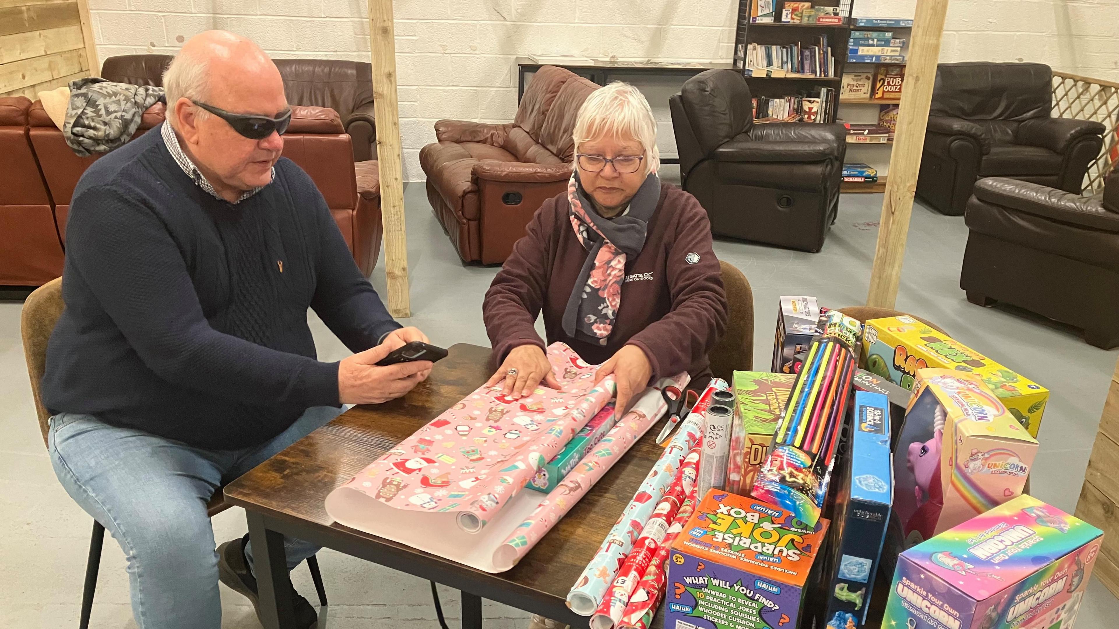 A couple are sitting at a table which has children's toys at one end such as a surprise joke box and unicorn toy and Colleen is wrapping a box with pink Christmas paper. 