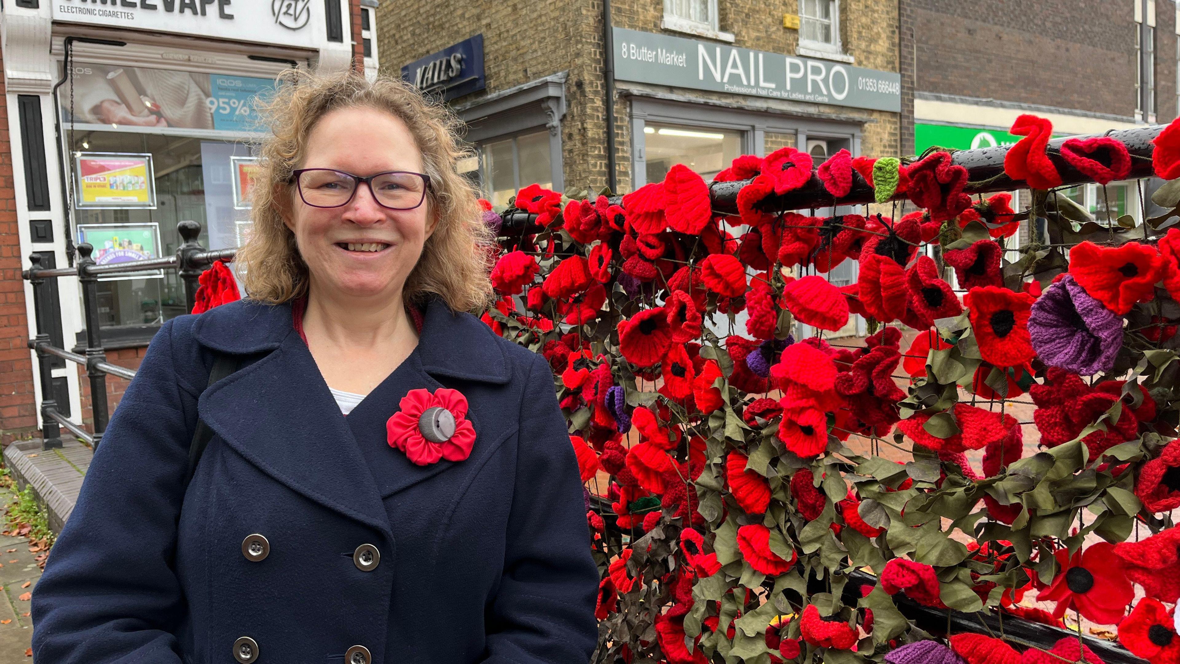 Sheila Willson is smiling and looking at the camera to the left of the photo. She is wearing glasses and a navy double-breasted coat with a large hand-made red poppy with a black button in the centre. Behind her is metal railing packed with red and purple knitted poppies, with artificial green leaves between them. In the background are shop fronts. 