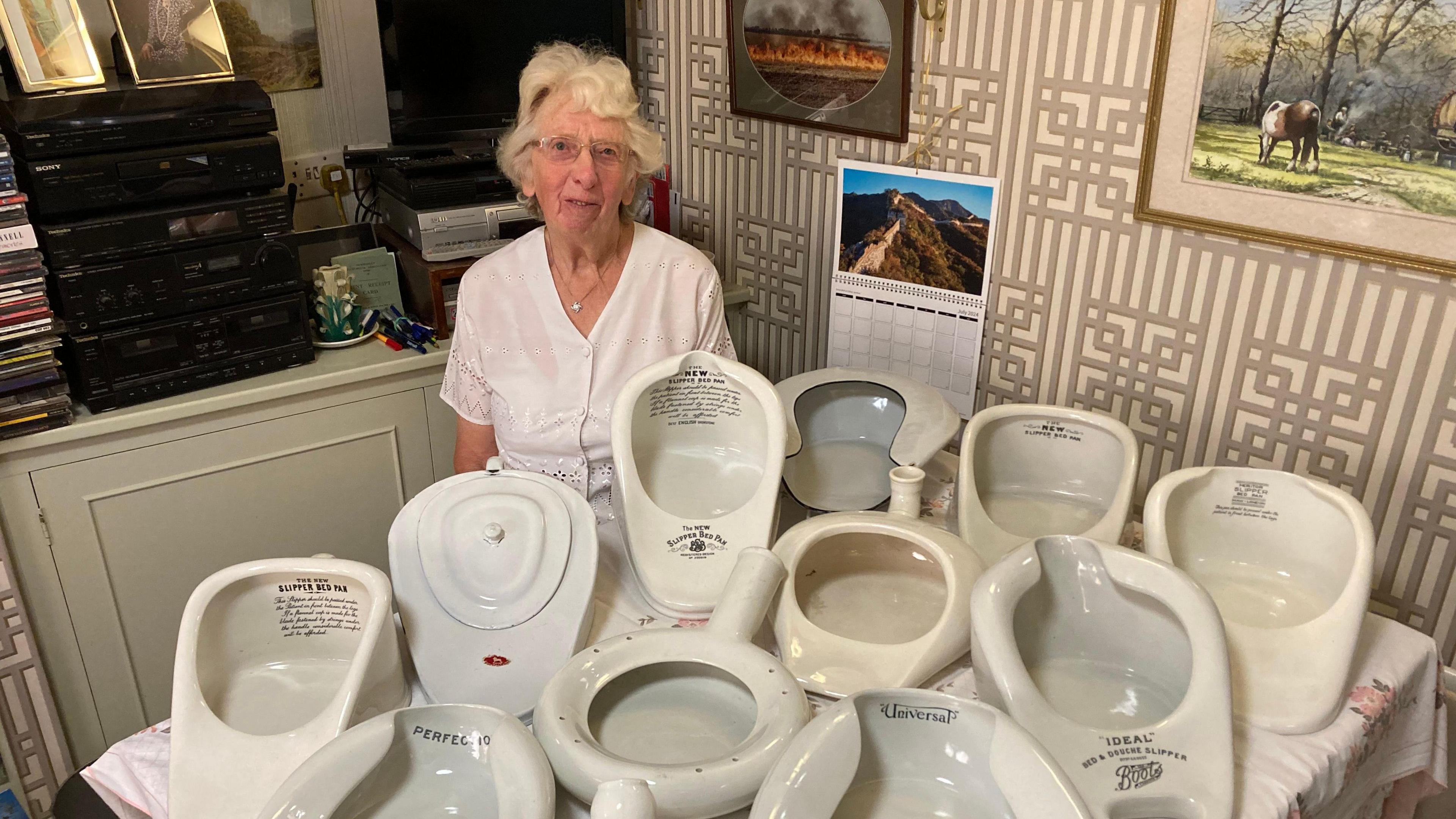 Mary Jacobs is pictured sitting behind some of her collection of white bedpans on a table. She is smiling, wearing a white top. The collection consists of an array of bedpans of all different shapes and sizes.