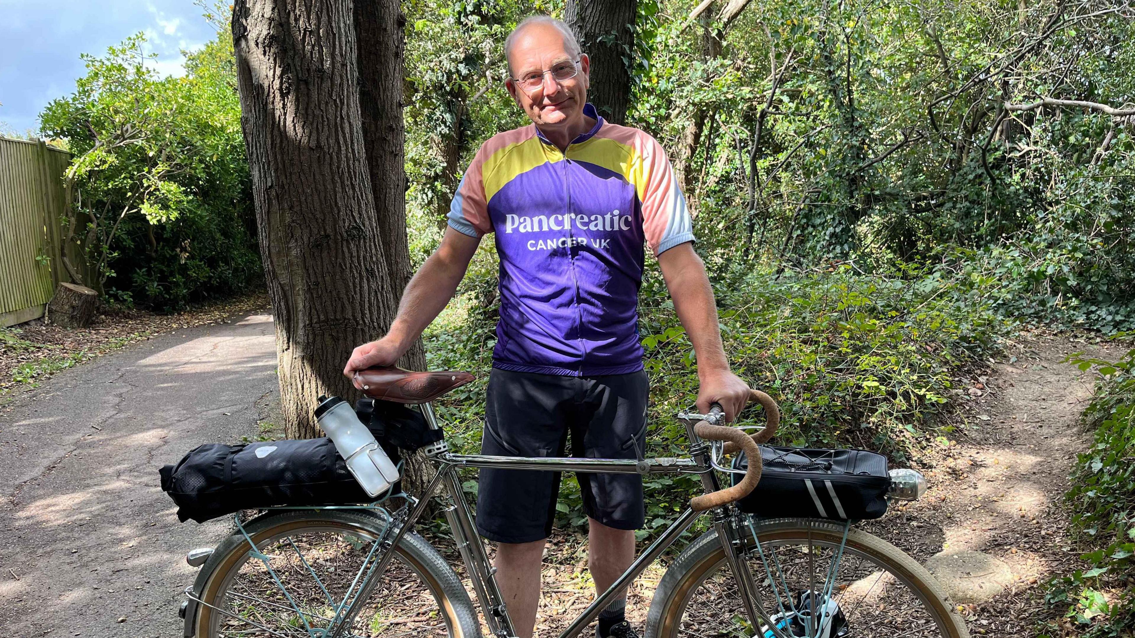 Steve Groves wearing a purple Pancreatic Cancer UK cycling top holding his chrome coloured 90 year old bicycle standing in a woodland area