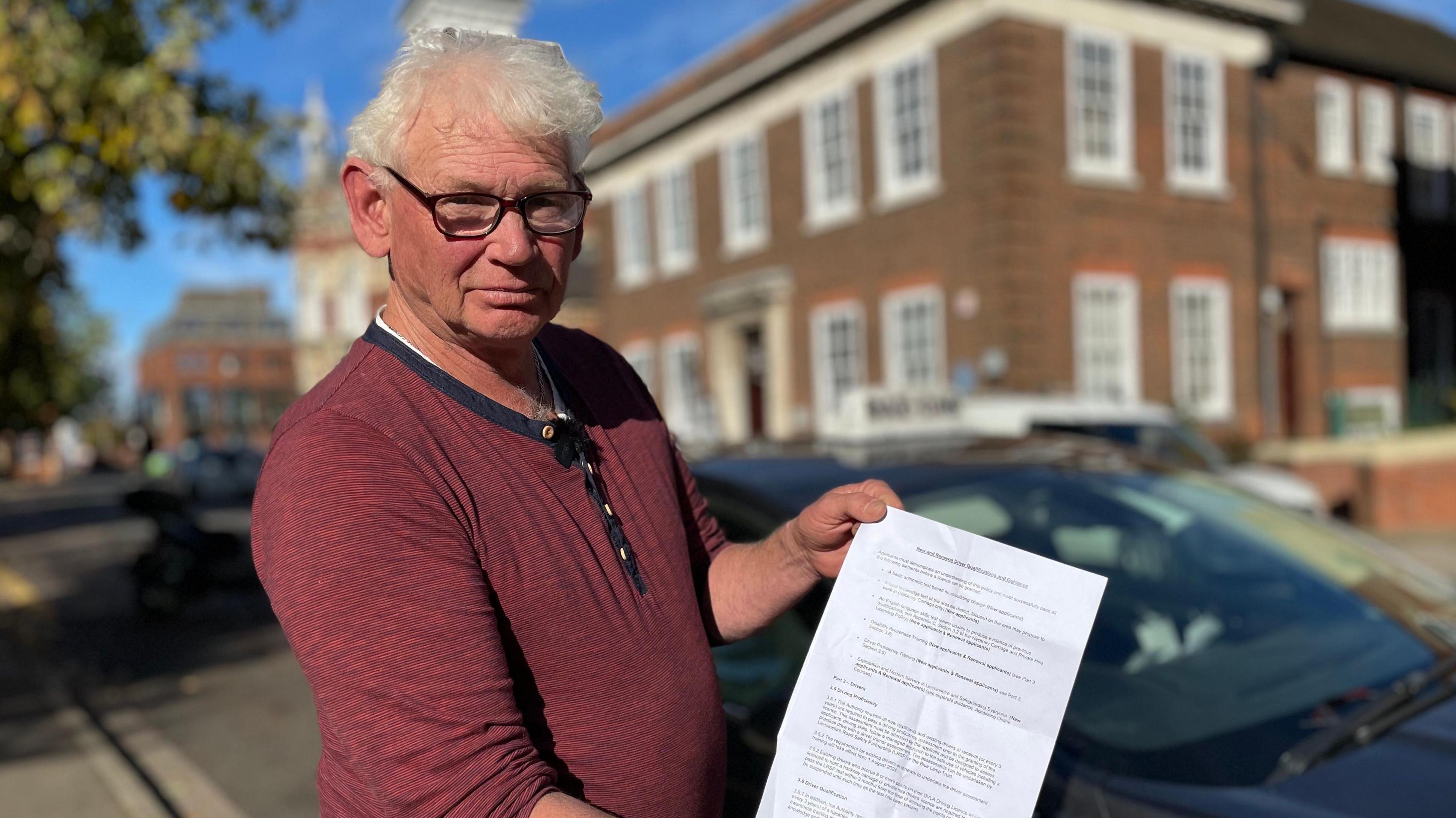 Barry Drake, wearing a red top, holds a print out of the renewal policy which says existing drivers must prove their English skills. A blurred council building and car are behind him.