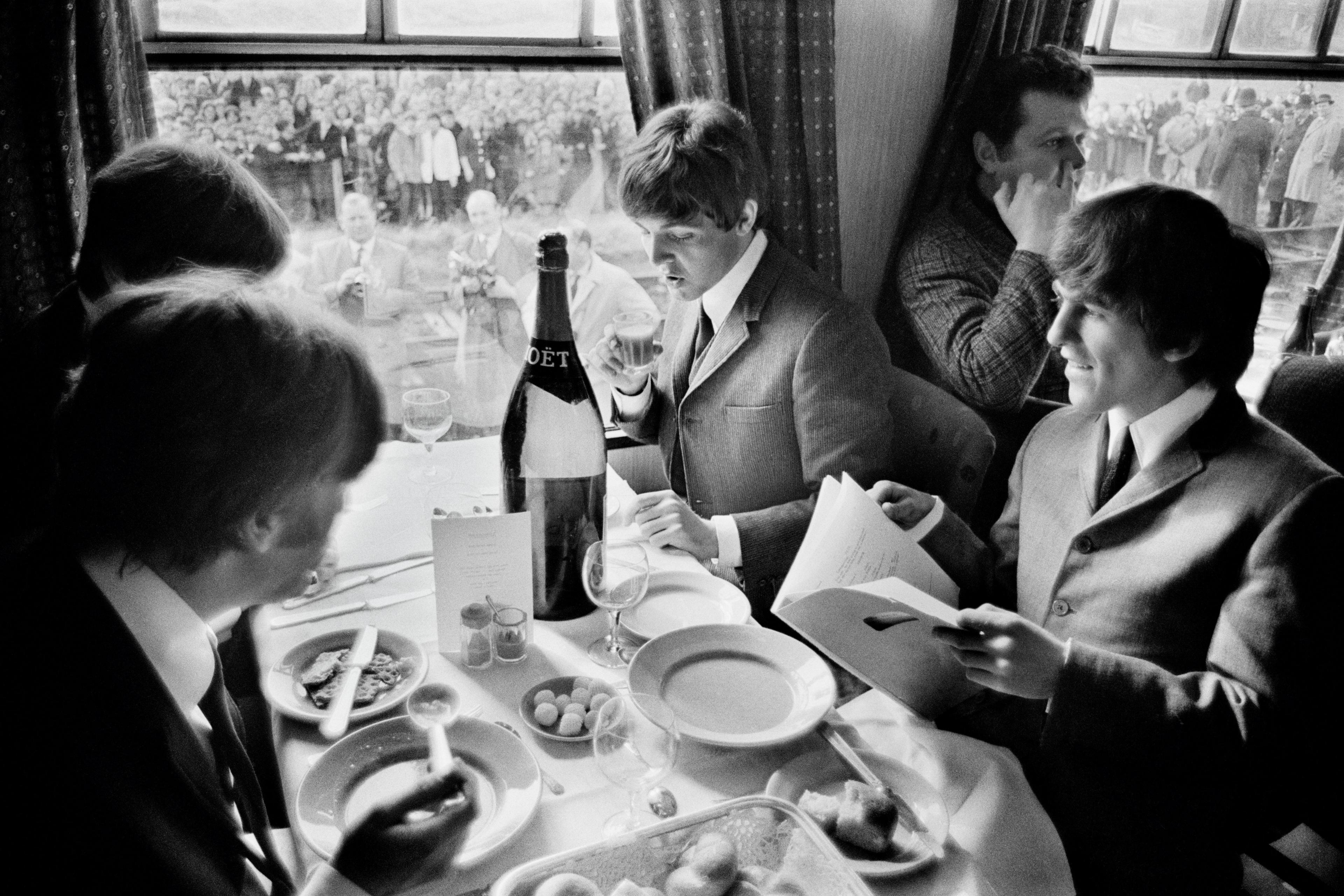 A black and white photo of John Lennon, Paul McCartney, George Harrison and Ringo Starr at Abbey Road Studios during the filming of A Hard Day's Night sitting on a train eating a meal off china plates. Crowds outside the train window look on.