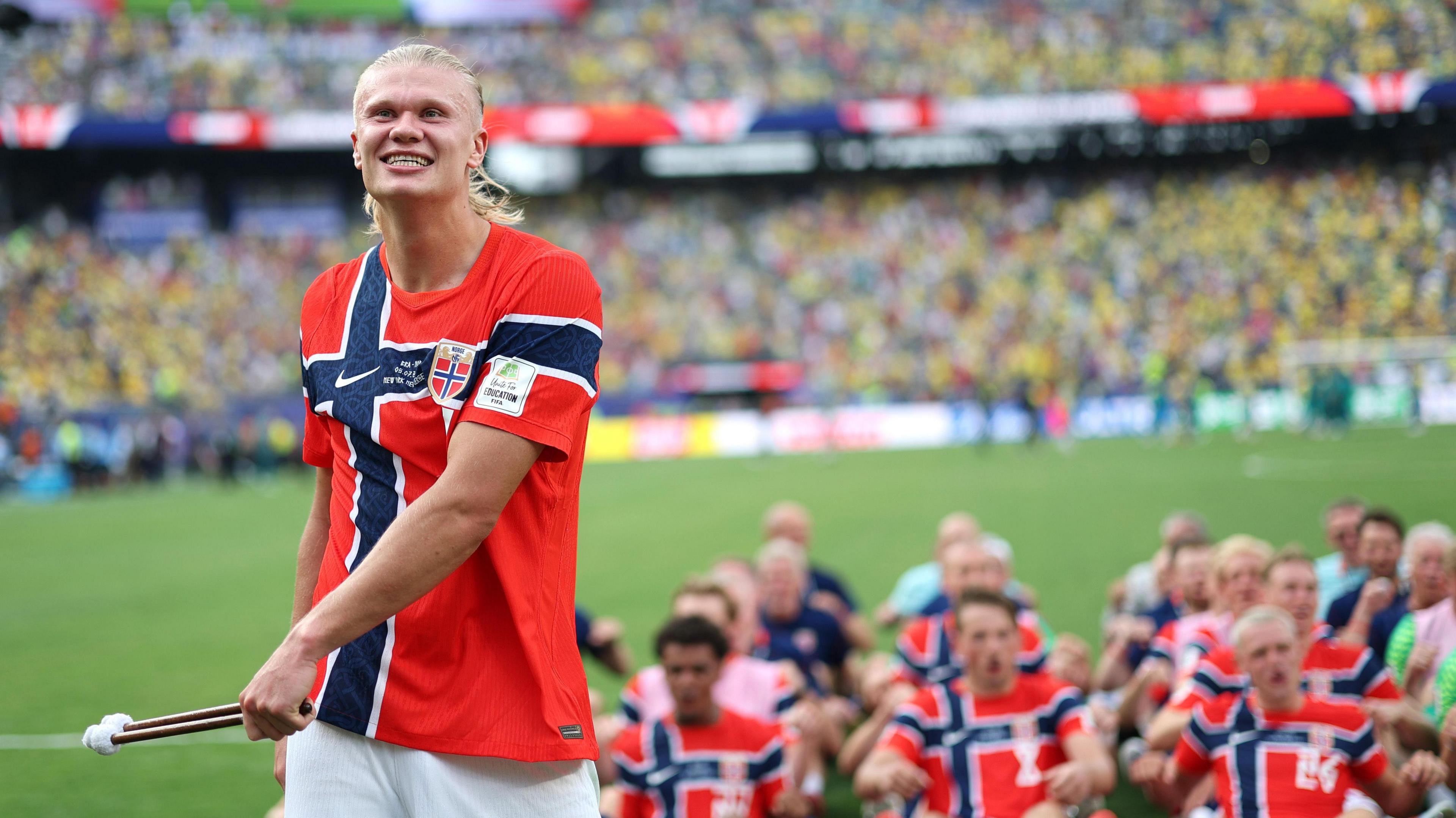 Haaland, wearing a red and blue Norwegian football jersey, smiles as he holds two drumsticks in his hand.