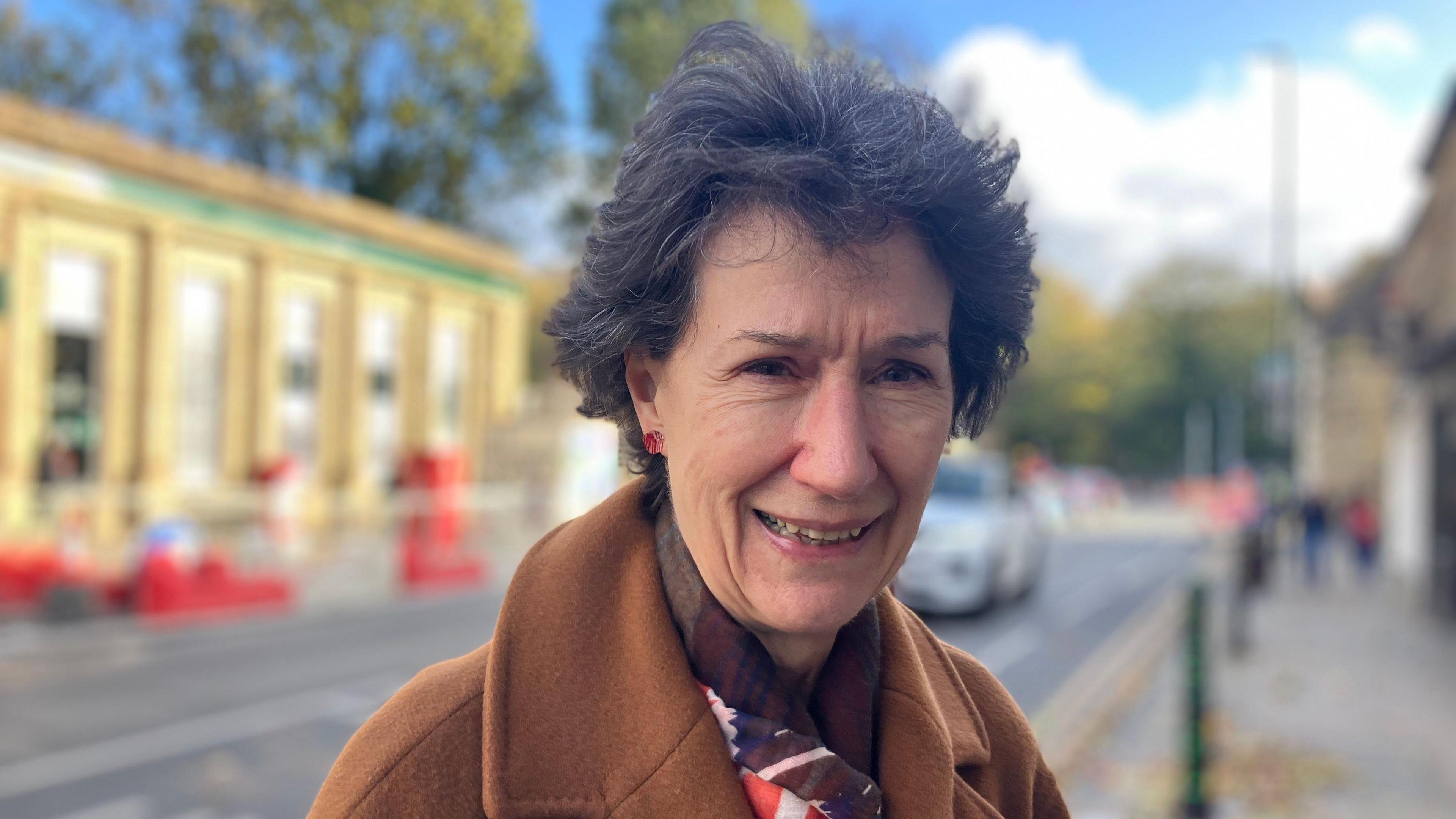 A woman with a brown coat stands on a pavement. A road is blurred behind her.