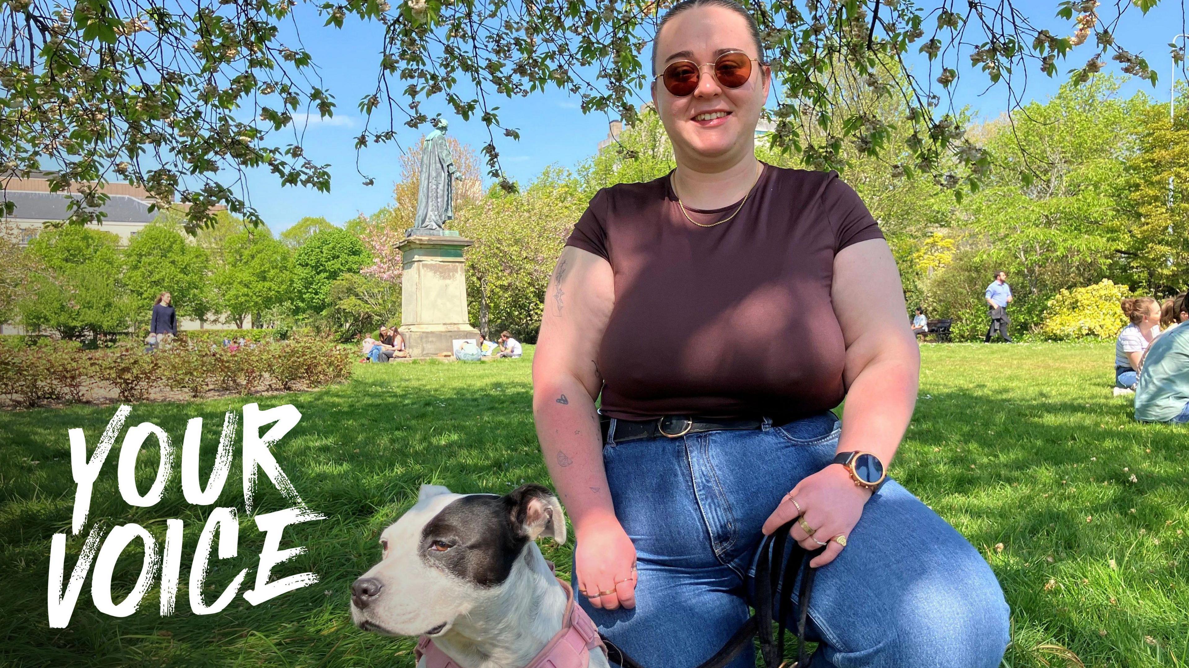 A young woman wearing a brown t shirt and jeans kneeling next to a black and white Staffordshire Bull Terrier in a pink harness in a sunny park.