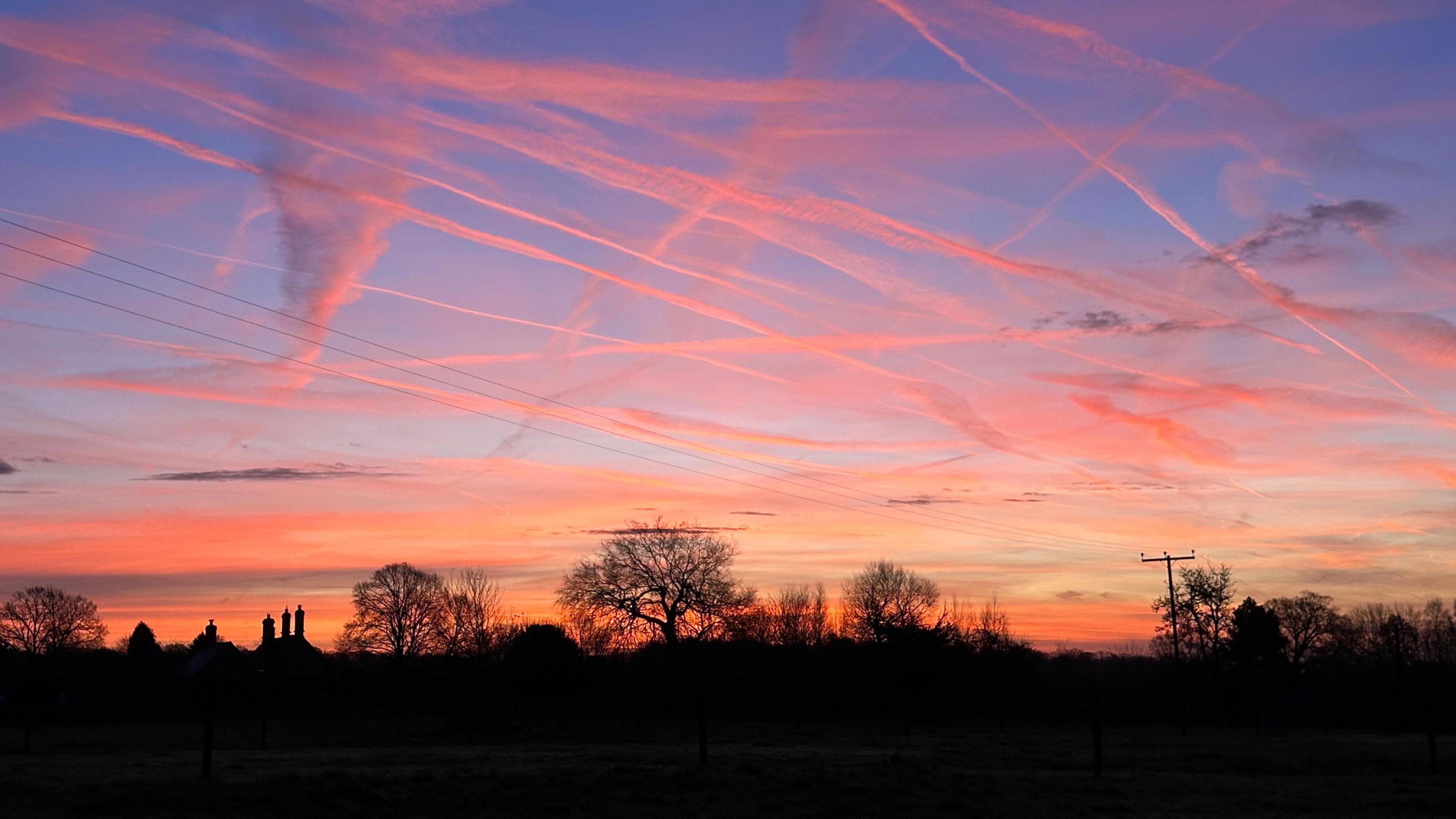 There are several trees and chimney tops silhouetted against the morning sky, which is turning blue but there is also a warm orange glow just above the horizon. Wisps of cloud shooting through the sky have also turned orange in colour.