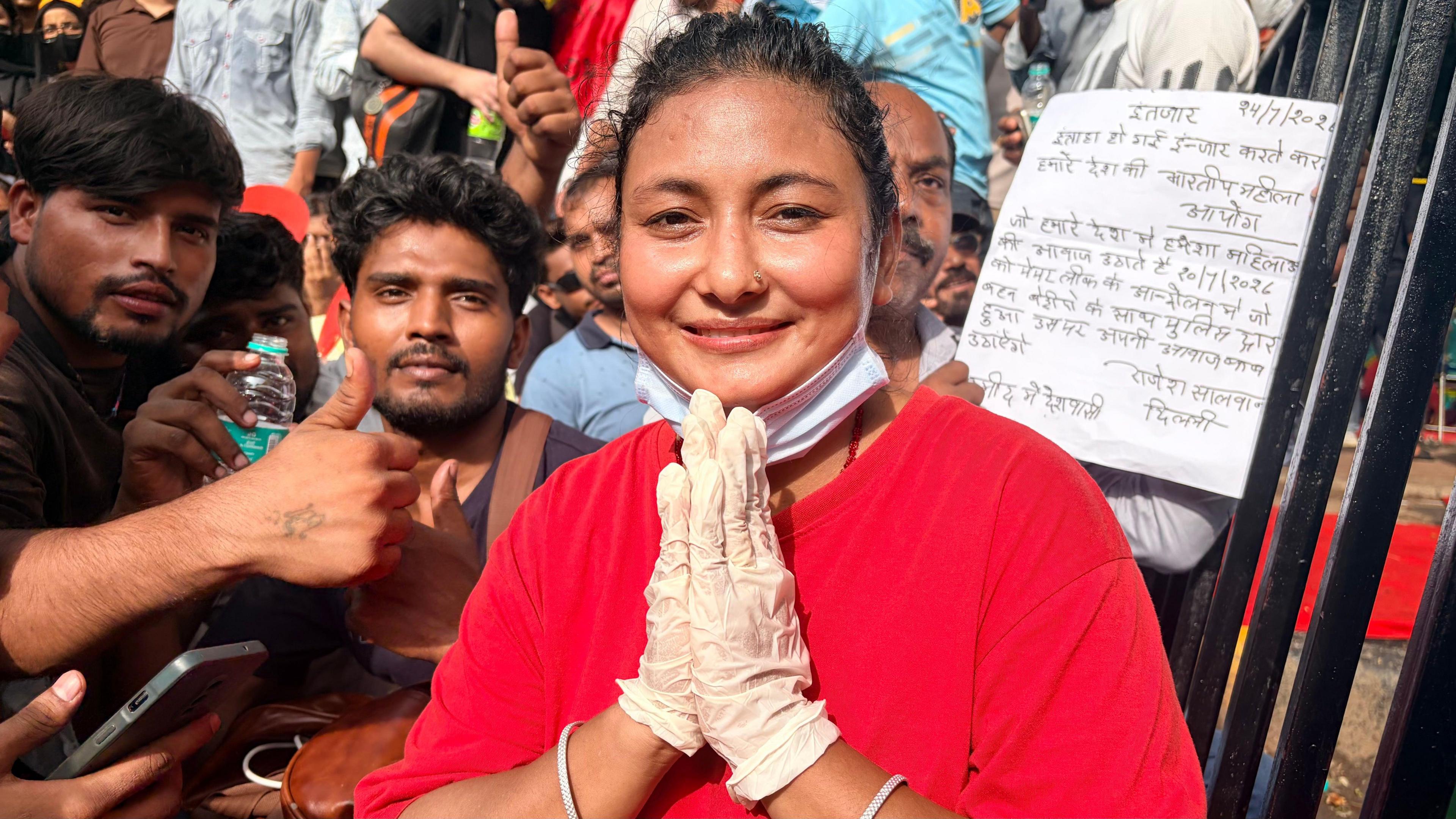 Radhika wears a bright red shirt, holding her hands as if saying a prayer and smiling, with people standing behind