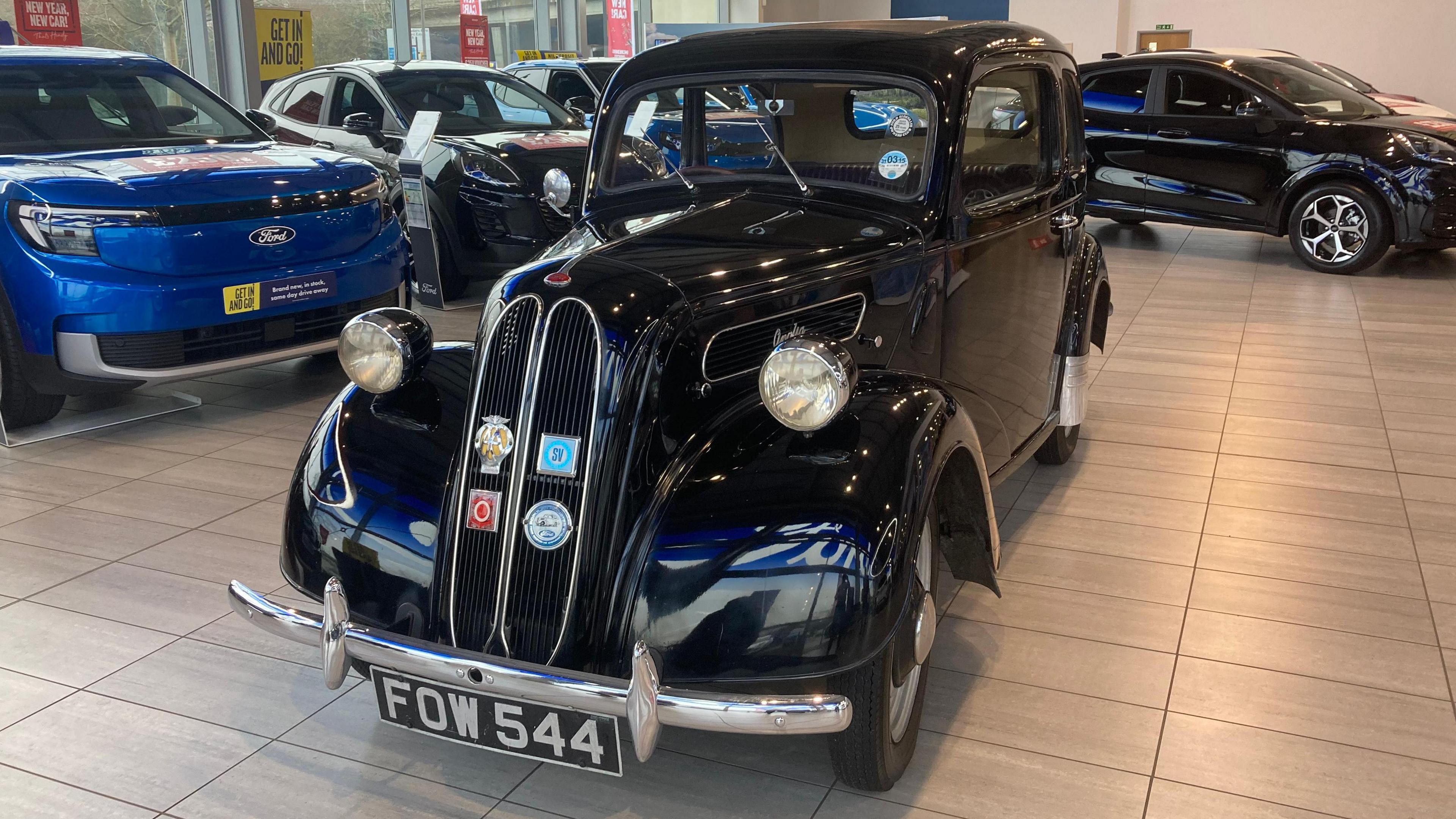 A vintage black Ford Anglia car in a showroom surrounded by modern Ford cars.