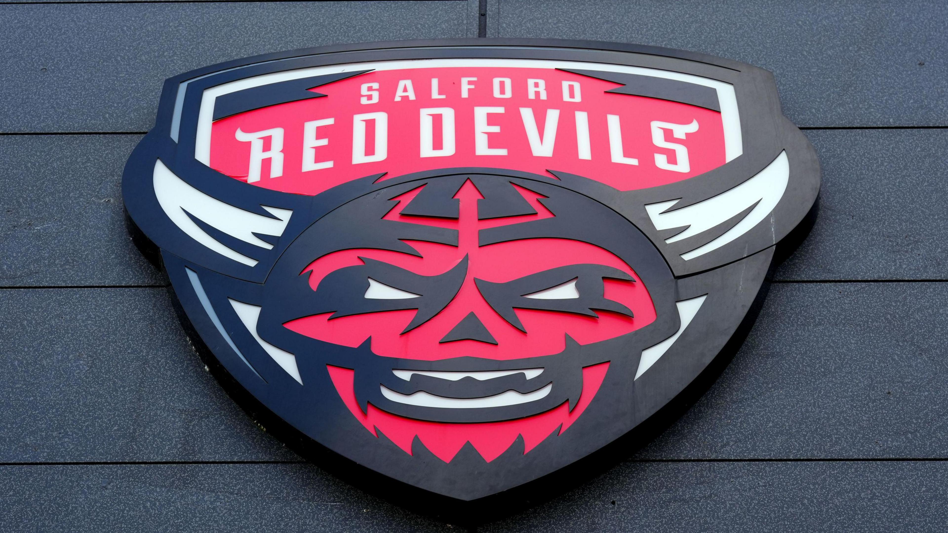 Salford Red Devils' logo mounted on a wall. It is a depiction of a white-horned, red-faced devil figure with white eyes.