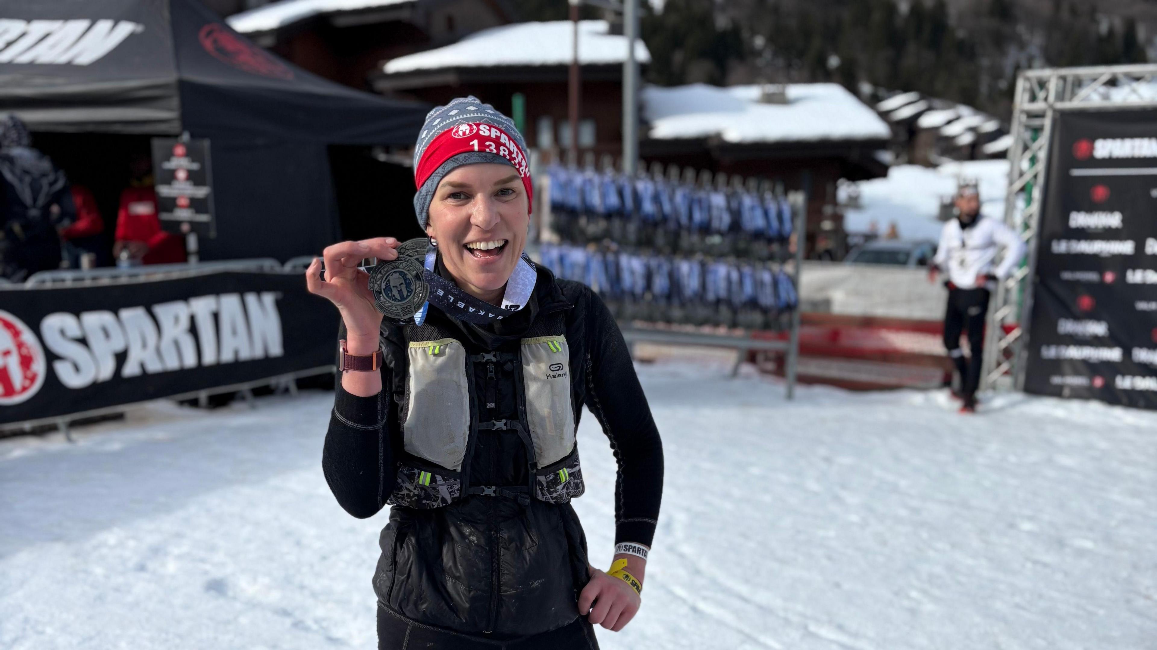 Fran Bullock holding a medal in her left hand and smiling at the camera. She is wearing a hat and there is snow around her. There is another person standing in the distance to the right. On the left is a black advertising sign with the words 'Spartan' in white.