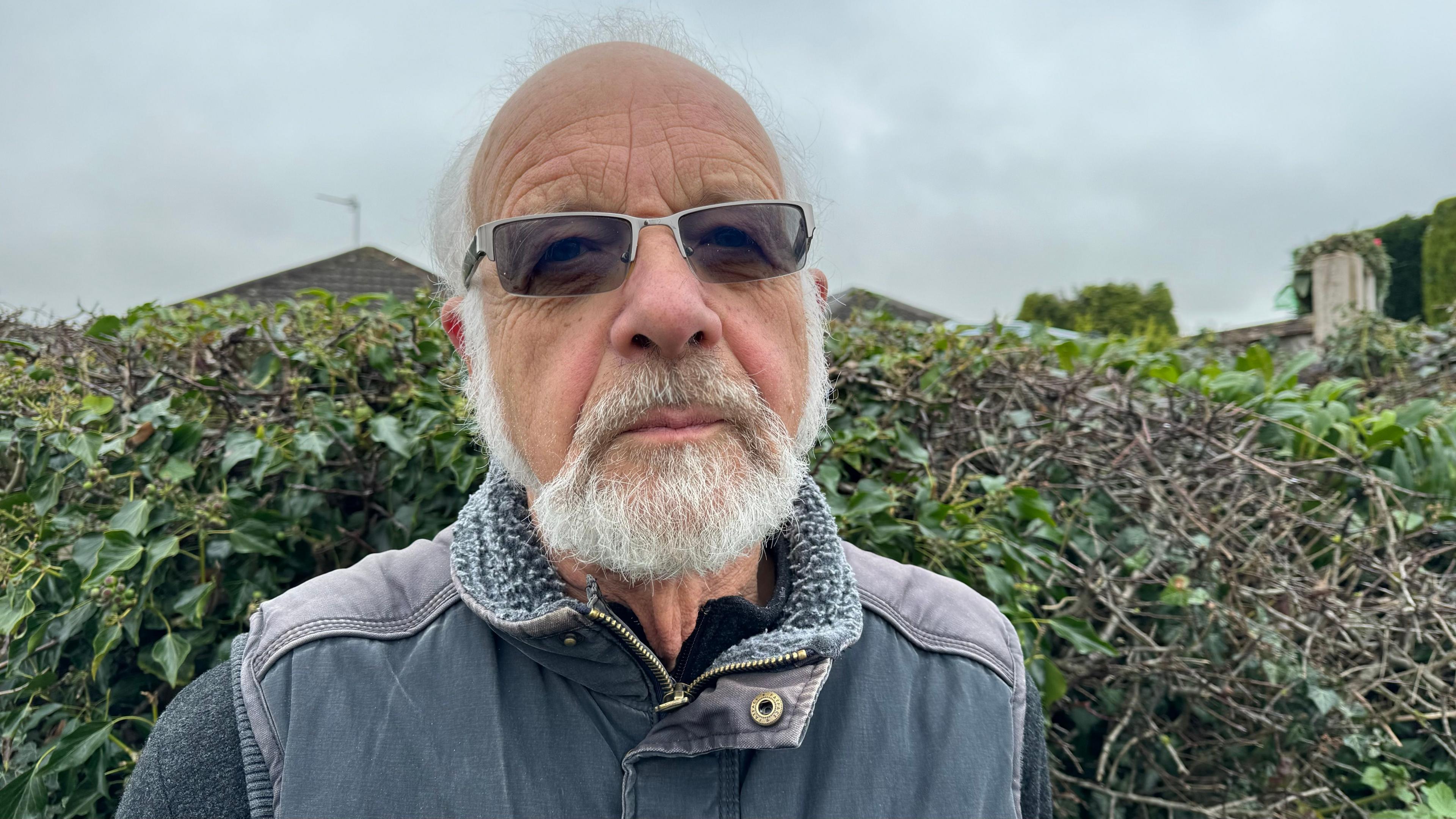 Robert Lowries from South Staffordshire standing in front of a hedge. He is wearing tinted glasses, a navy gillet and a navy jumper 