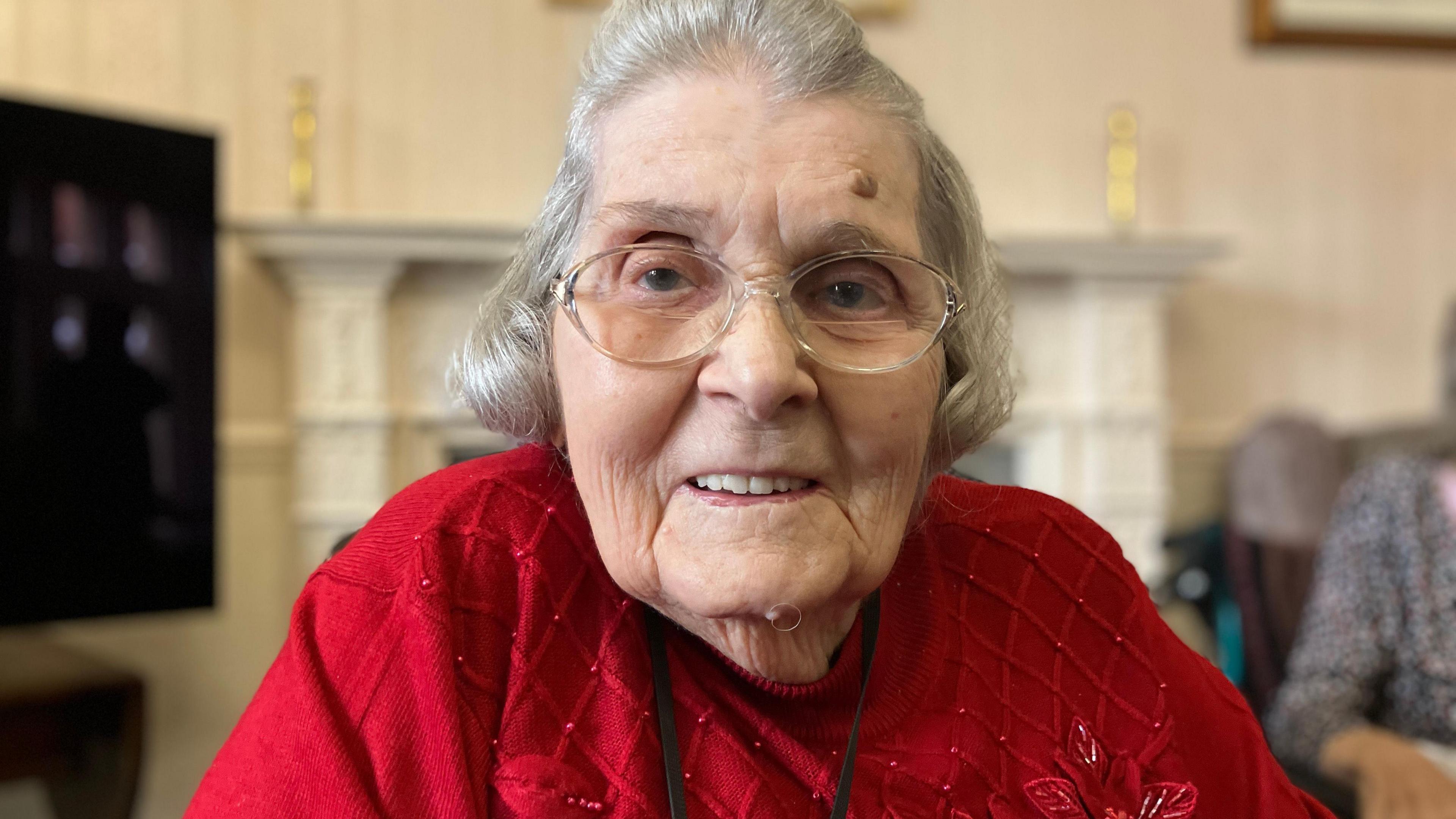 Janet Wills is sitting in her care home's front room. She is wearing a red jumper and glasses. She is smiling.