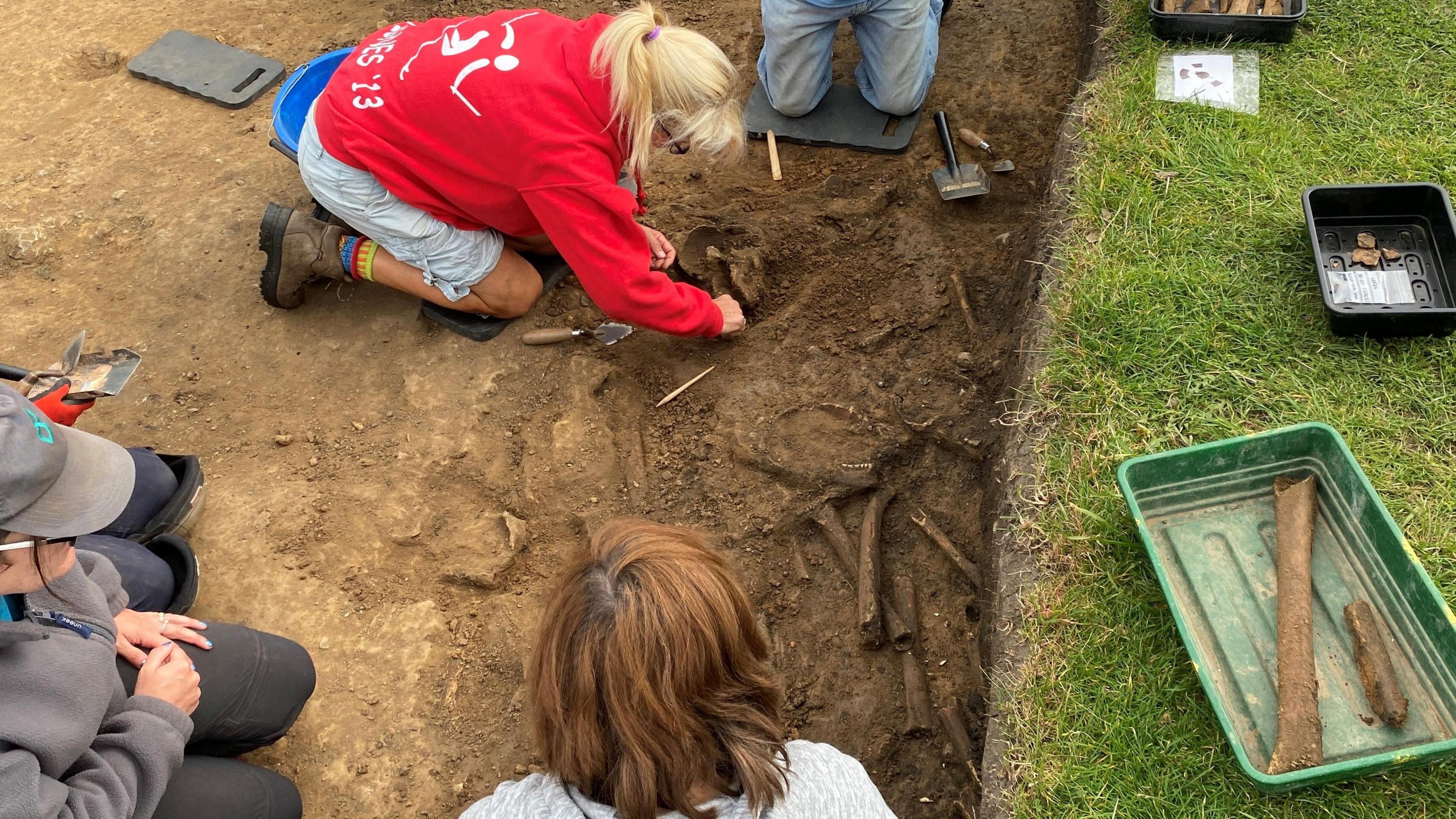 A woman wearing a red top is on her hand and knees scraping dirt from a skull while three other people kneel in the trench. There are other bones visible.