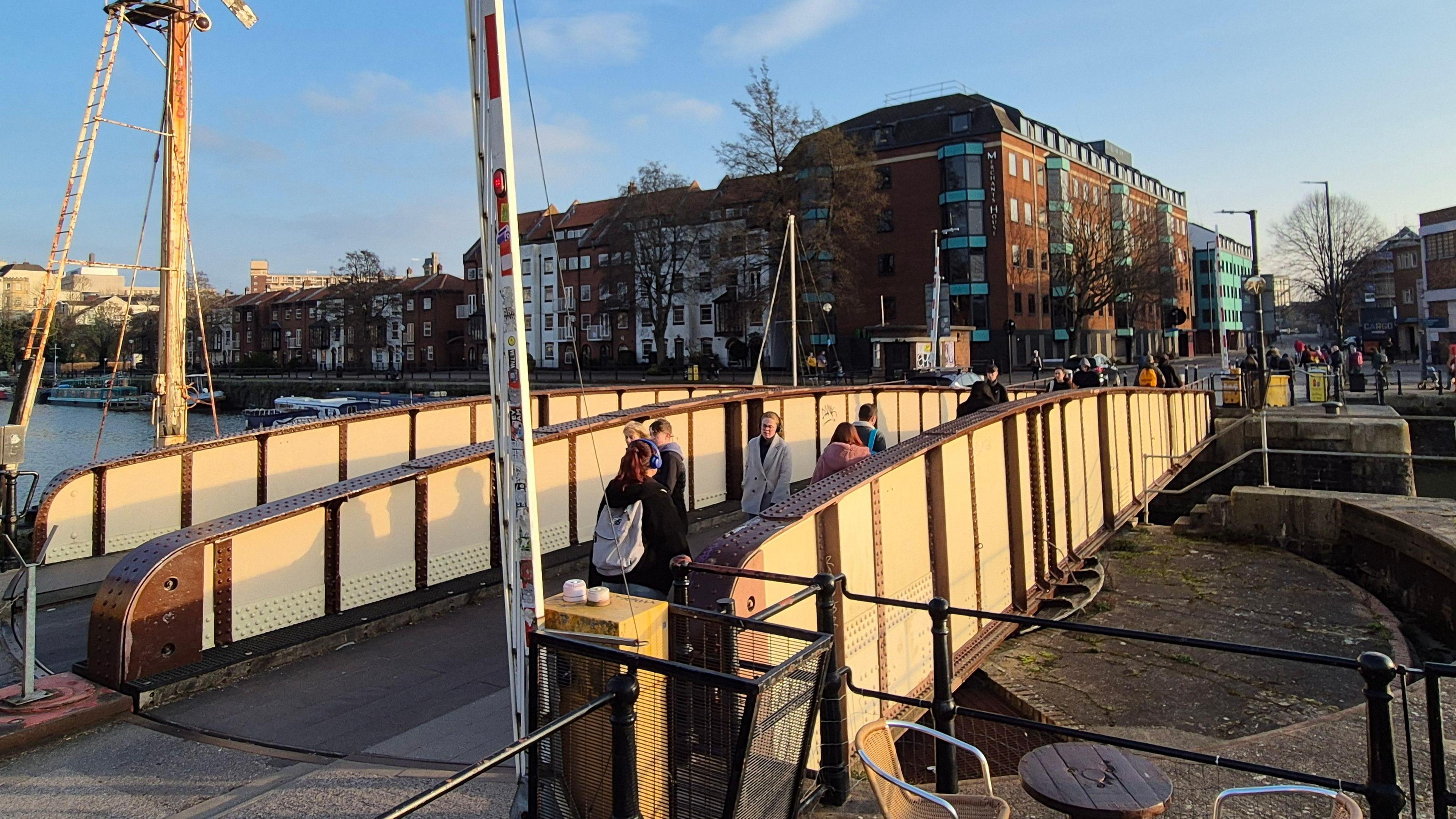 Prince Street Bridge in Bristol. It has metal barriers on either side and people are walking across it. In the background are residential houses.