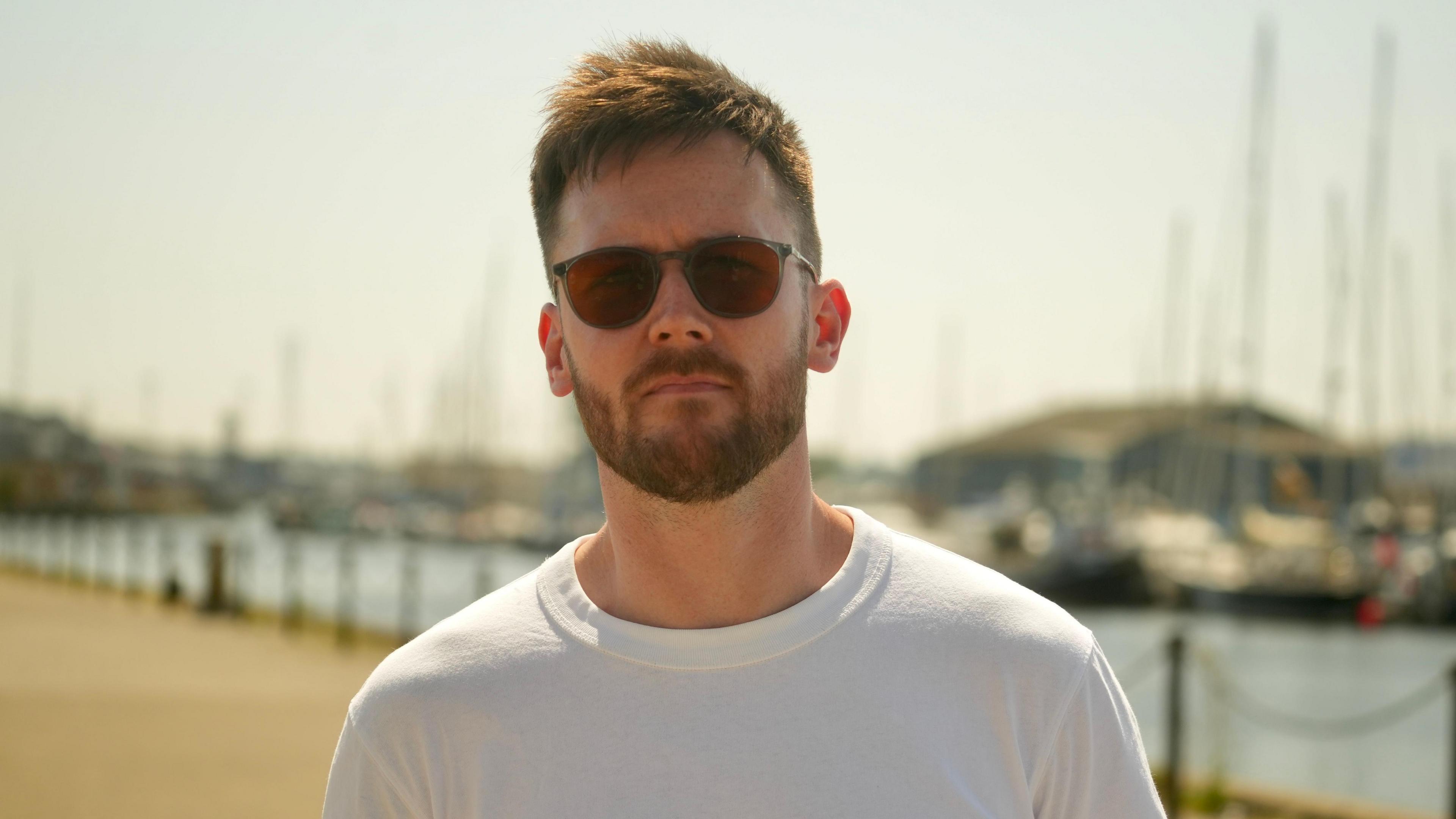 Tom Griffin standing in front of boats and a dock, looking into the camera. He has brown hair, rounded glasses and a beard. He is wearing a white t-shirt.
