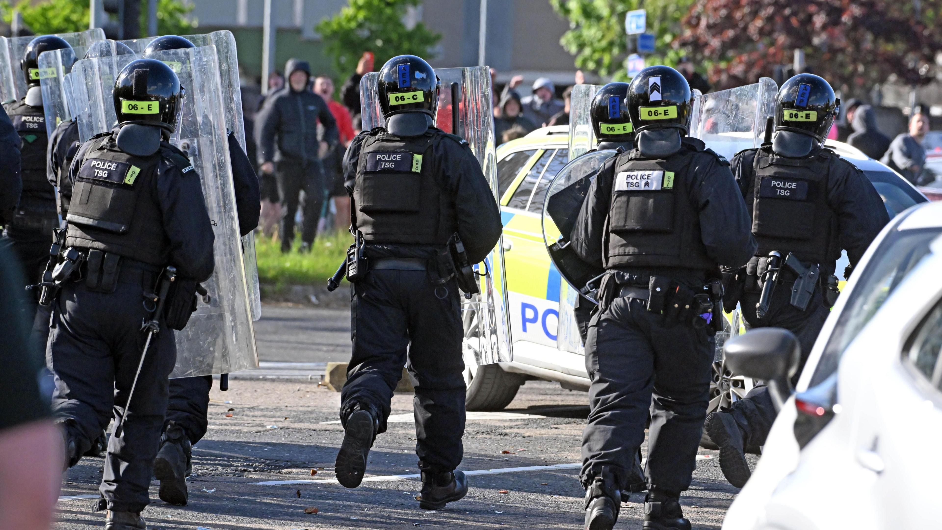A shot of five police officers in black riot gear and holding large plastic shields running towards a crowd in the distance. There is a marked police car parked in between the officers and the crowd.