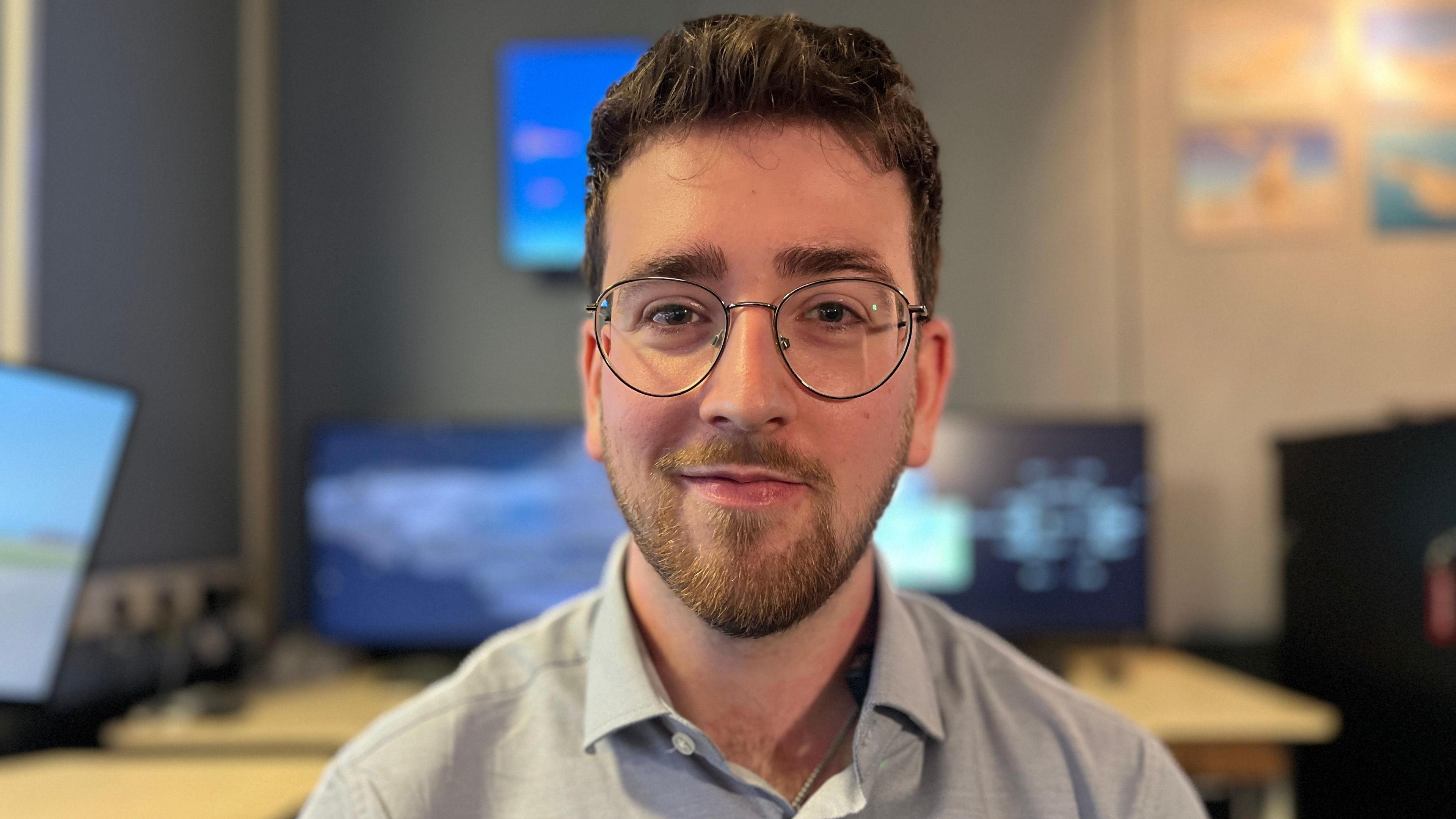 Lewis Solomon stands in front of computer screens. He is wearing glasses and a blue shirt, and looking directly at the camera. 