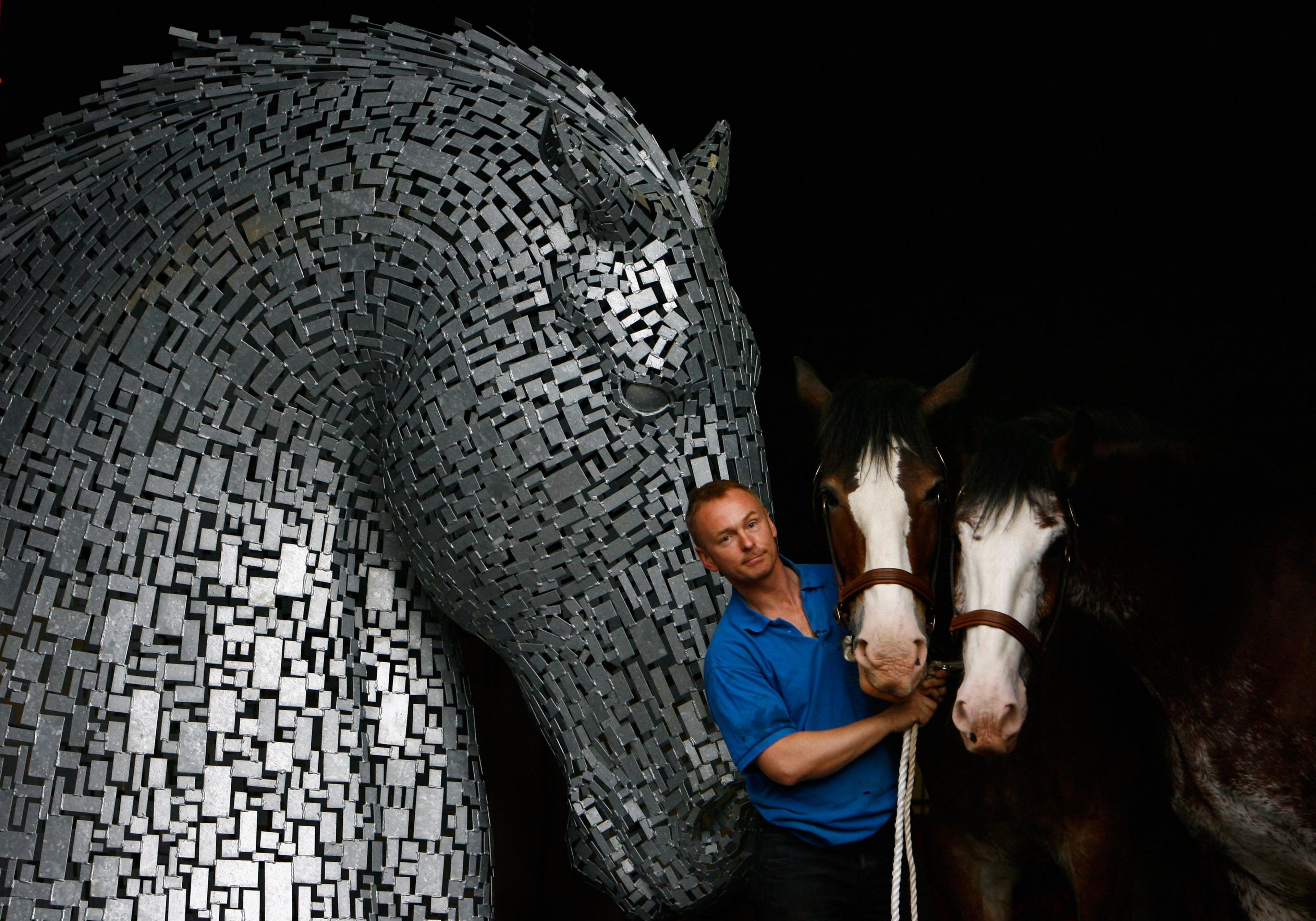 Sculptor Andy Scott wearing a blue t-shirt and holding the reins of two Clydesdale horses, stands next to one of his sculptures of the Kelpies