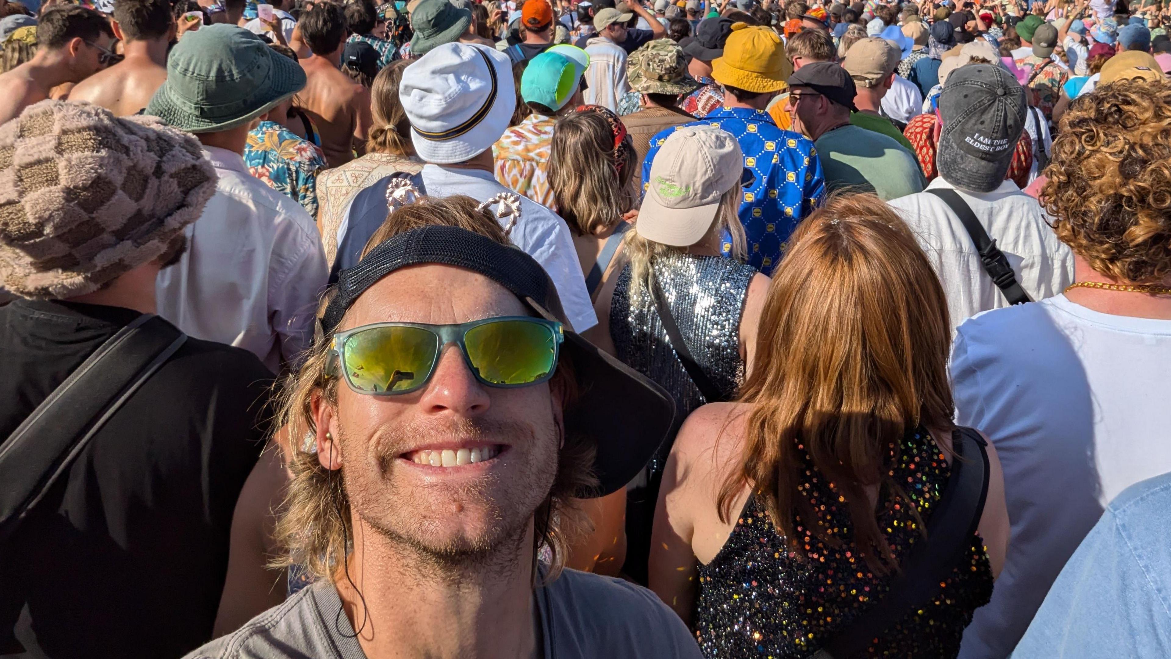 Lewis Simonds, smiling at the camera, with a cap on his head, to the side, sunglasses on, in front of a large crowd of people, at Glastonbury