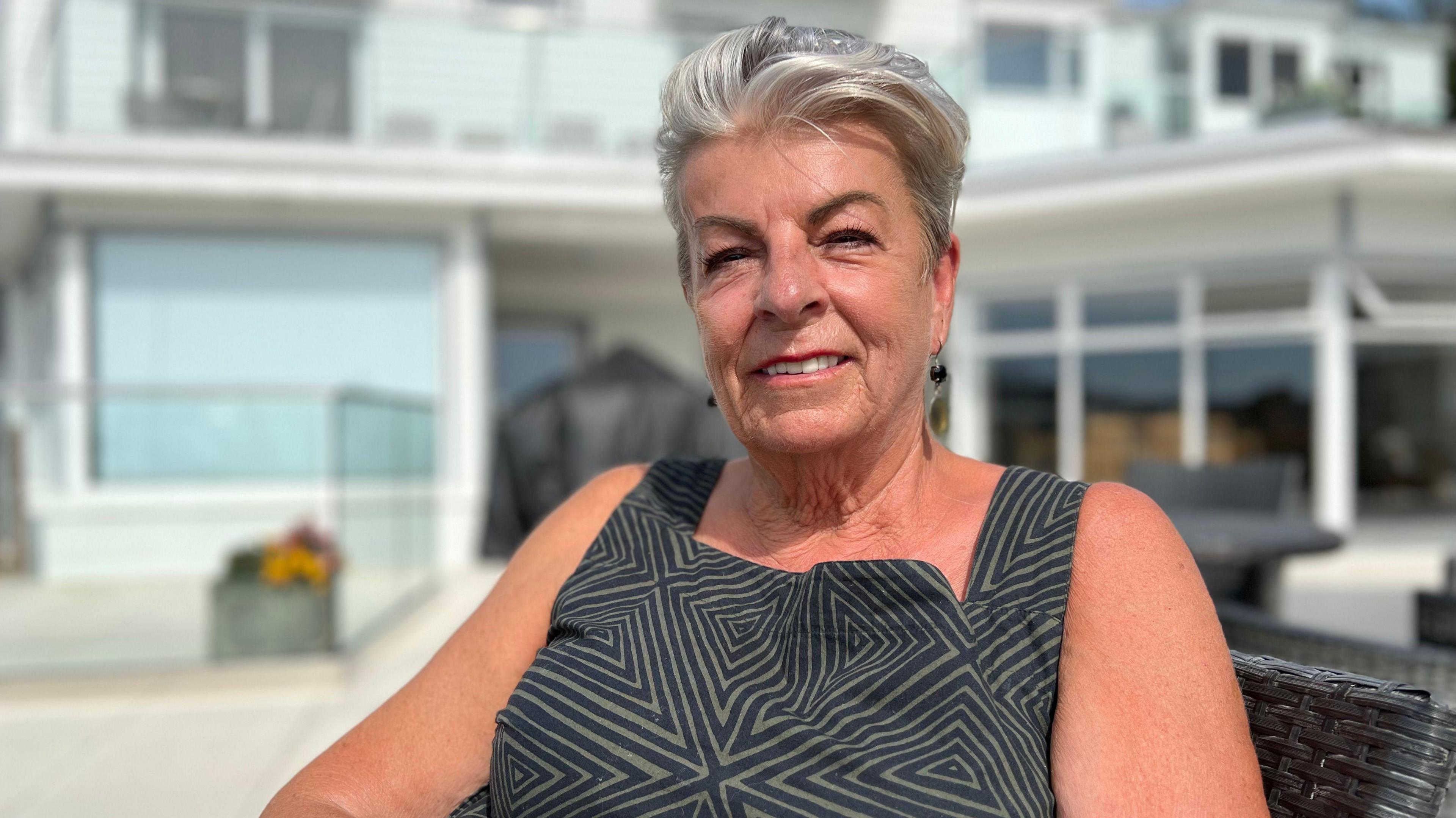 Penelope Bromley has short white/grey hair and is wearing black dangly earrings, a black and brown square-neck top with thick straps and a triangle pattern. She is smiling at the camera and the building behind her is blurred.