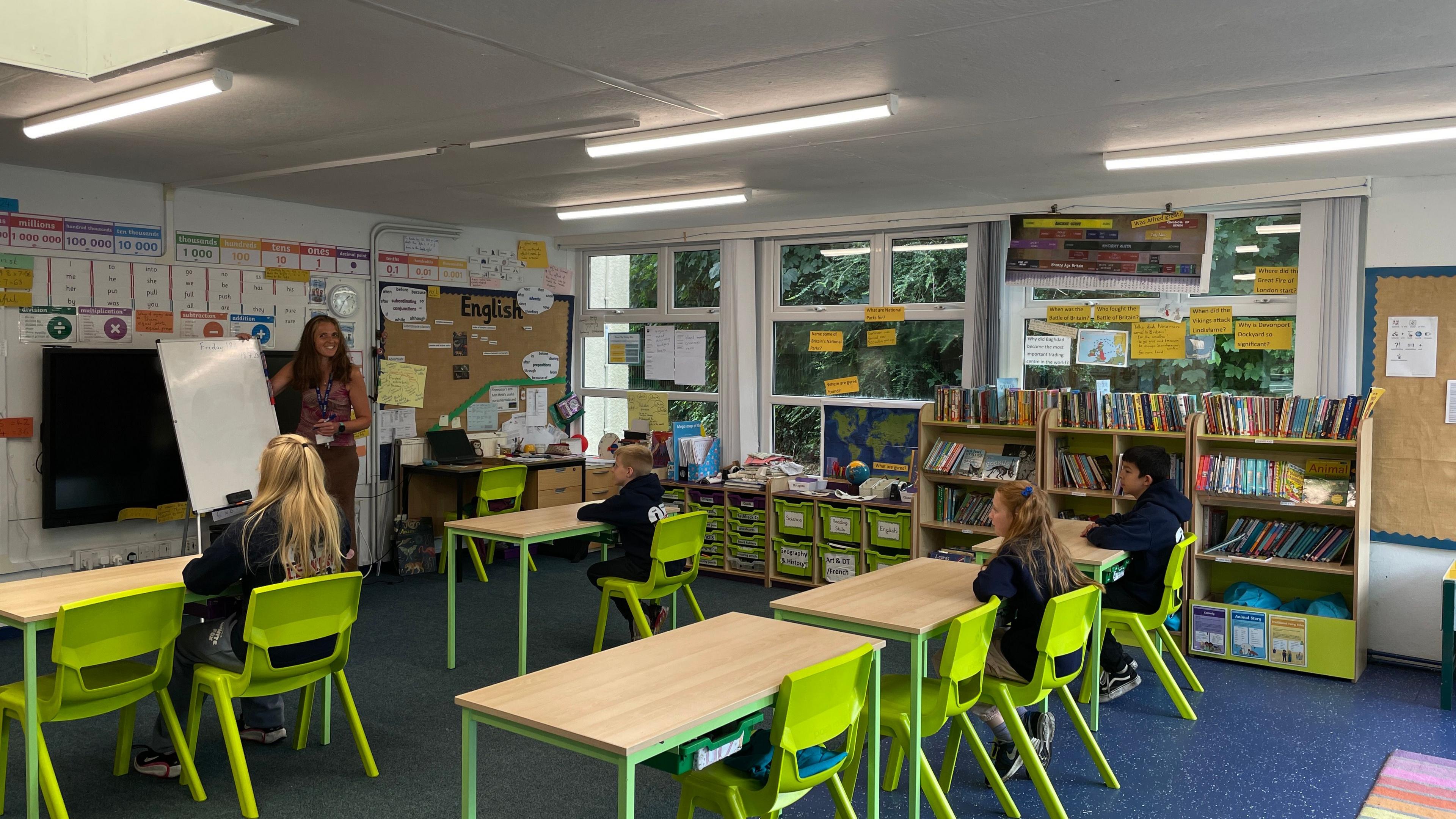 A classroom. It has five wooden tables with green table legs and green chairs. There are four students, facing away from the camera towards the front of the classroom. There is a female teacher at the front next to a whiteboard.