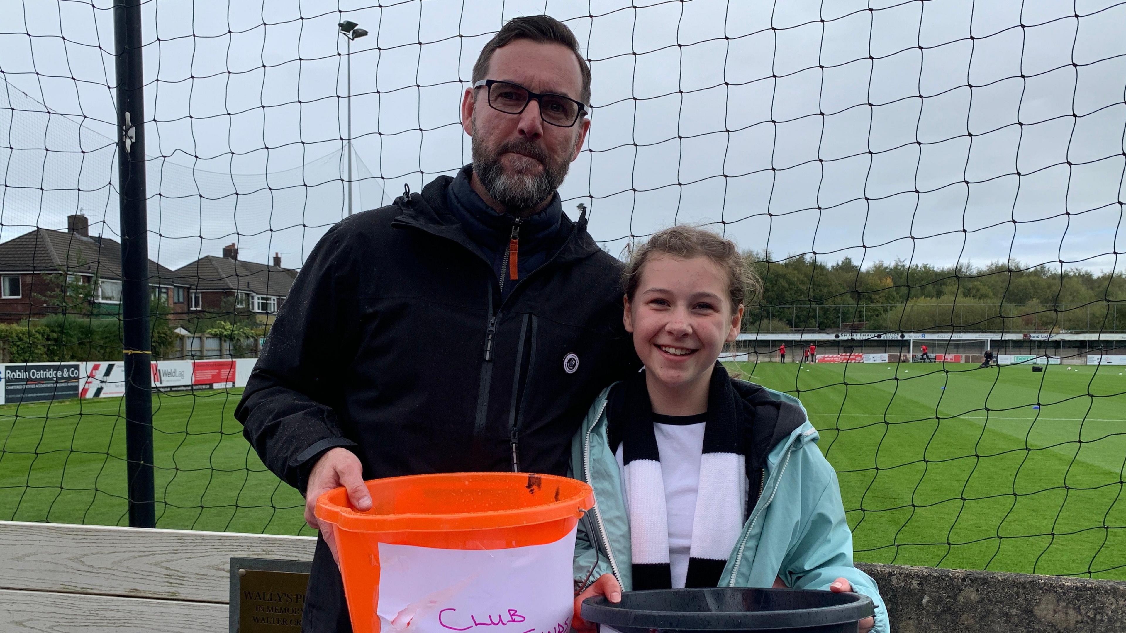 Father and daughter holding fundraising buckets with pitch behind them