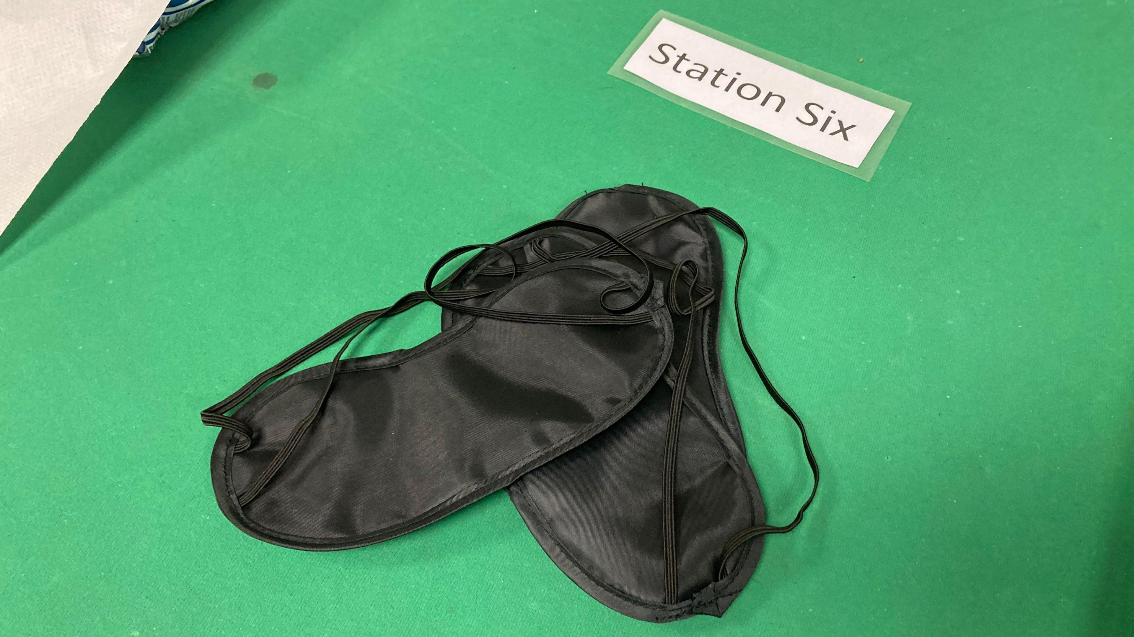 Two black eye masks sit on a table that is covered in a green felt fabric. 