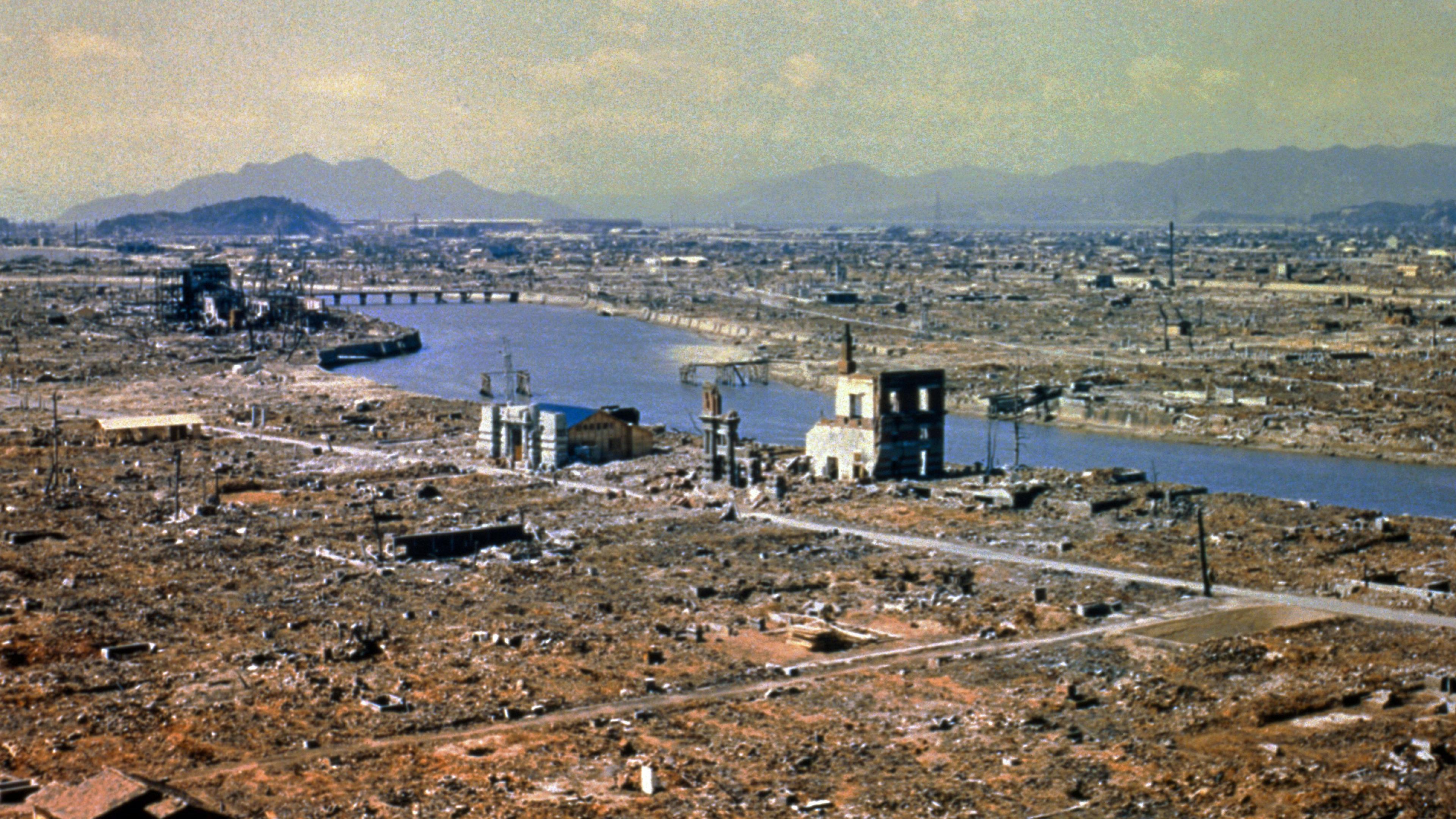 The destruction caused by the nuclear explosion over Hiroshima. The landscape is largely flattened with visible debris from properties. The shells of some properties near a wide river as visible. The ground looks brown.