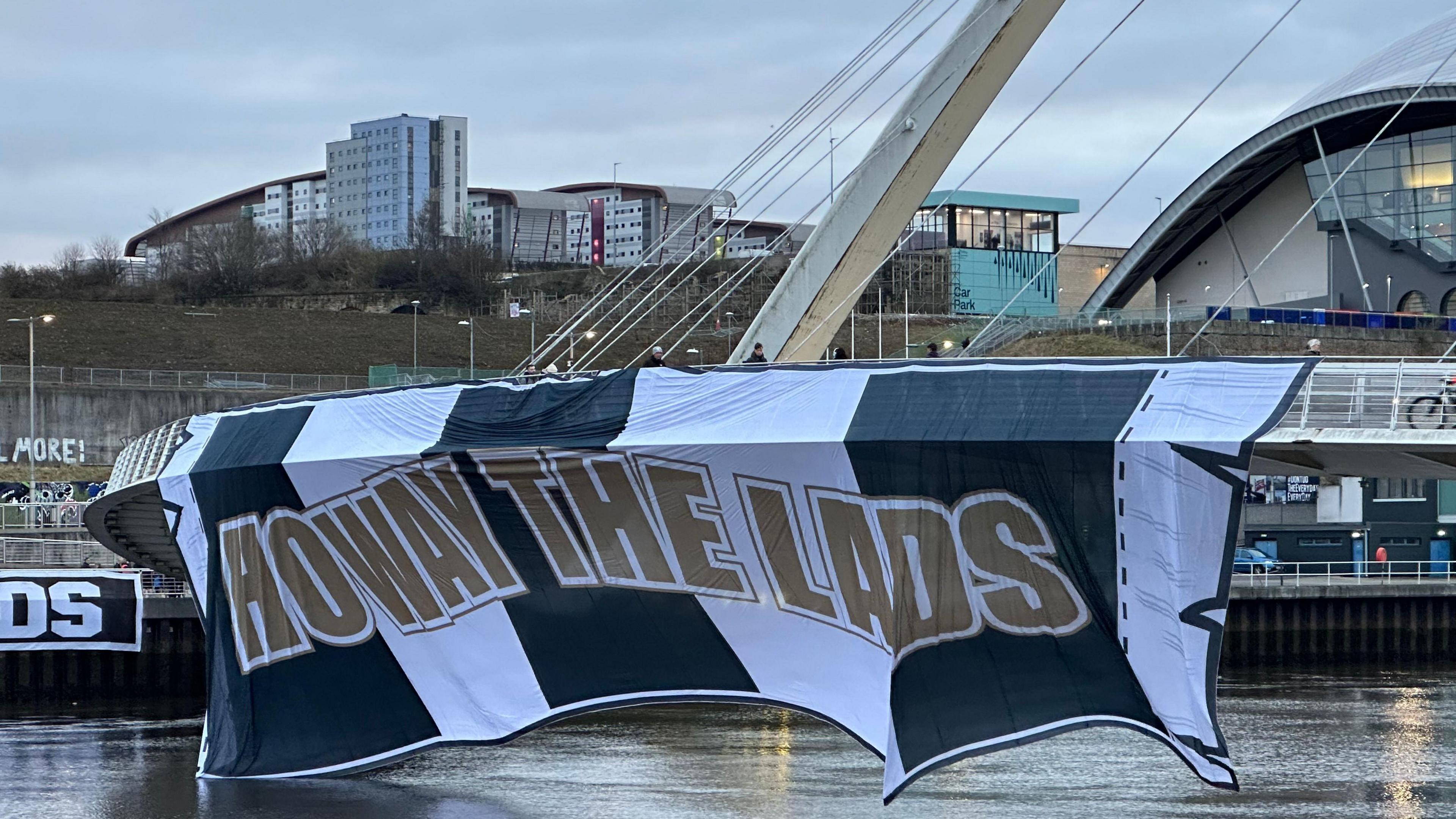 A big black and white flag which reads 'Howay the Lads' hanging off the side of the Millennium Bridge.