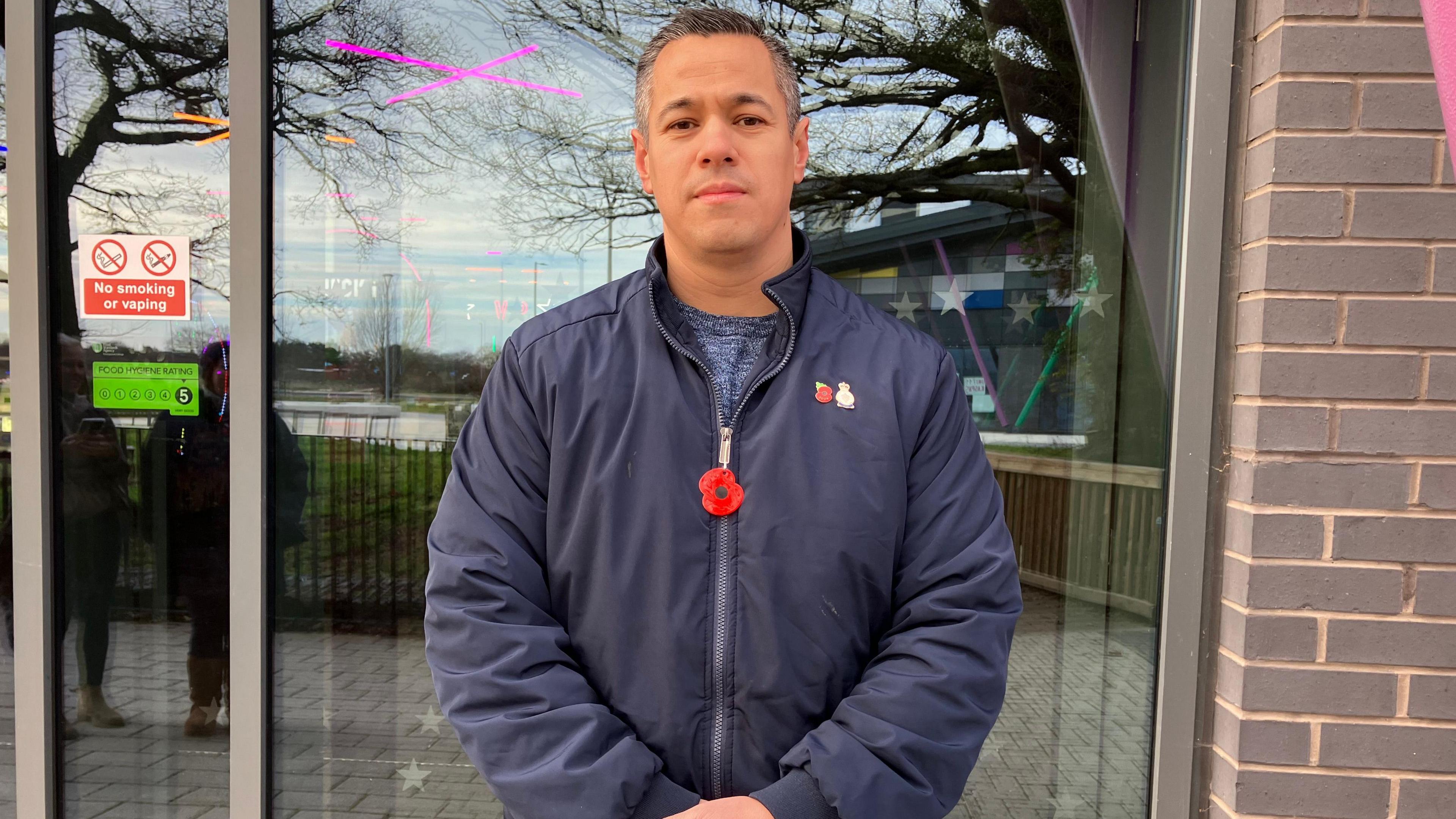 Freddie Bulpit stands in front of a shop. He is wearing a blue bomber jacket that is zipped up and a blue jumper. There is a poppy on his jacket. He has short, grey hair.