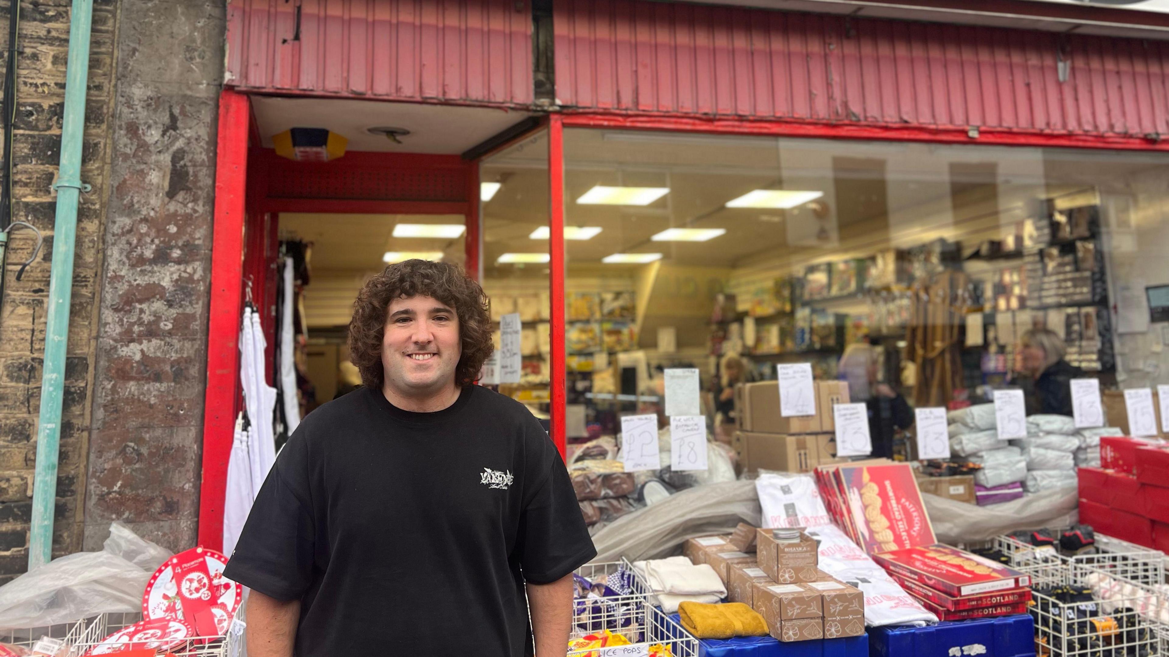 A man in a black t-shirt stands in front of a store called 'Dons'. Lots of stock sits outside the shop alongside him