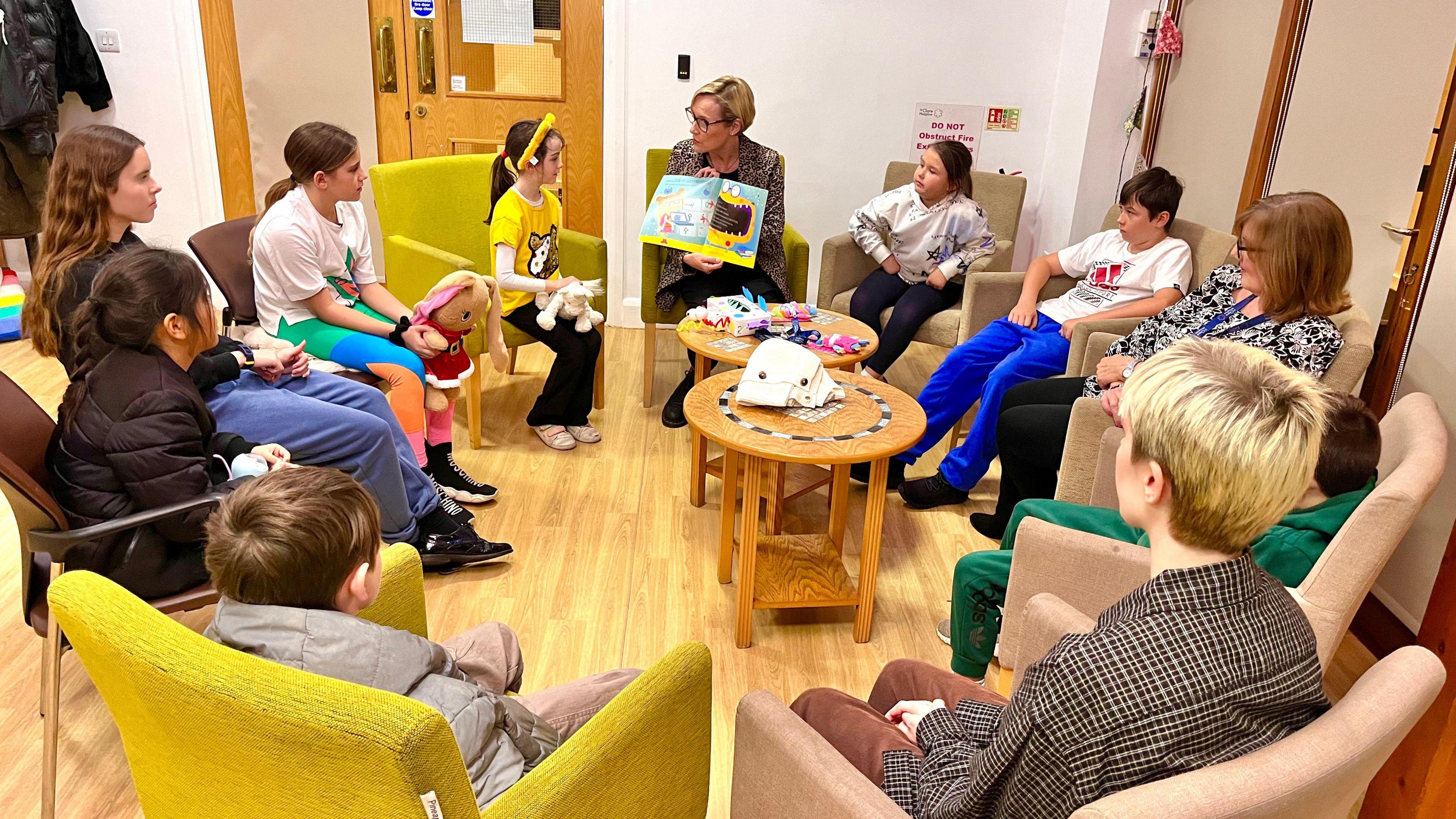 A group of children and adults sit around a table and look at a picture book. The room is brightly lit and the children look engaged. 