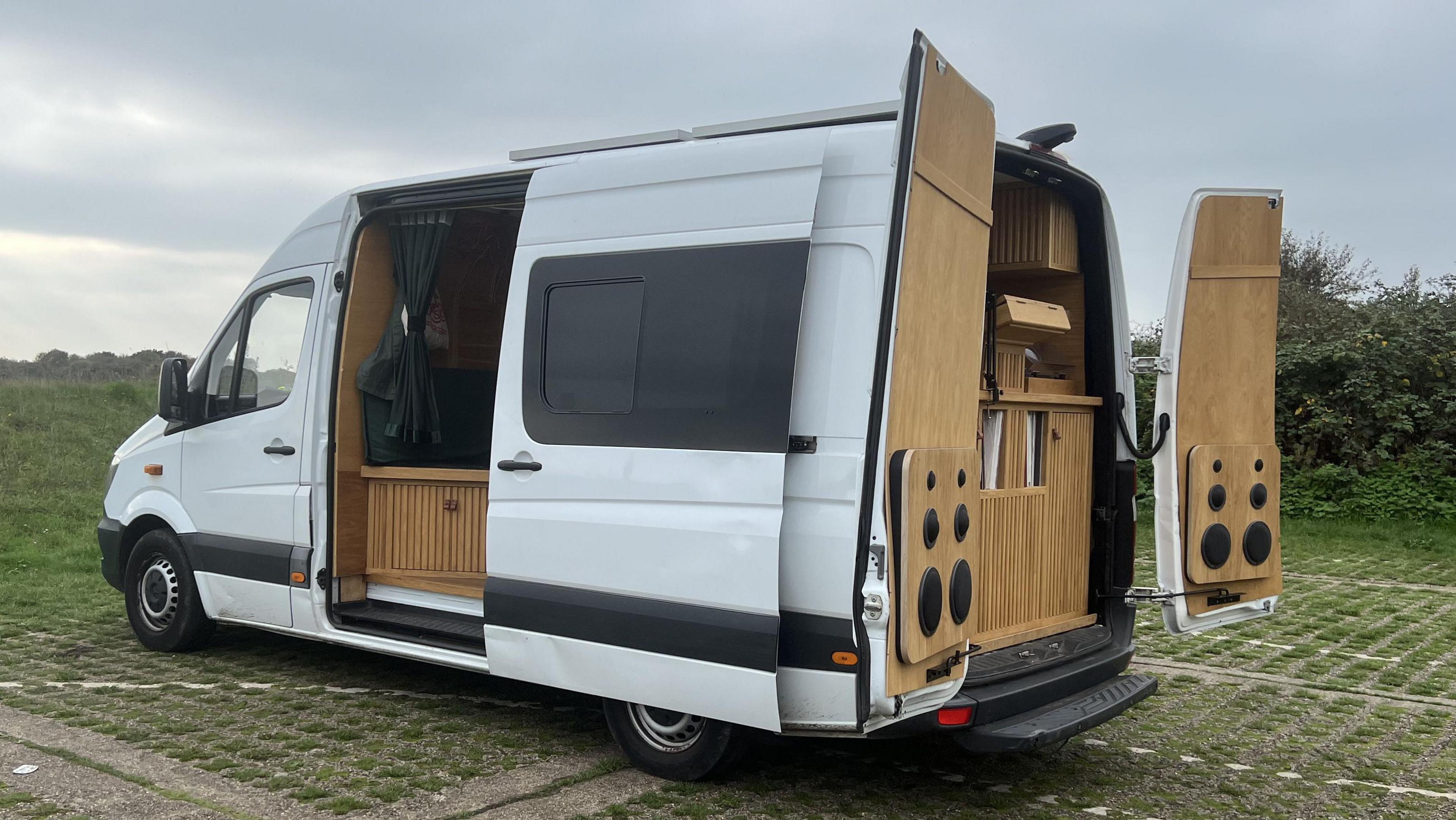 The outside of the white van parked in a green field. The doors are open showcasing the interior woodwork and speakers. 