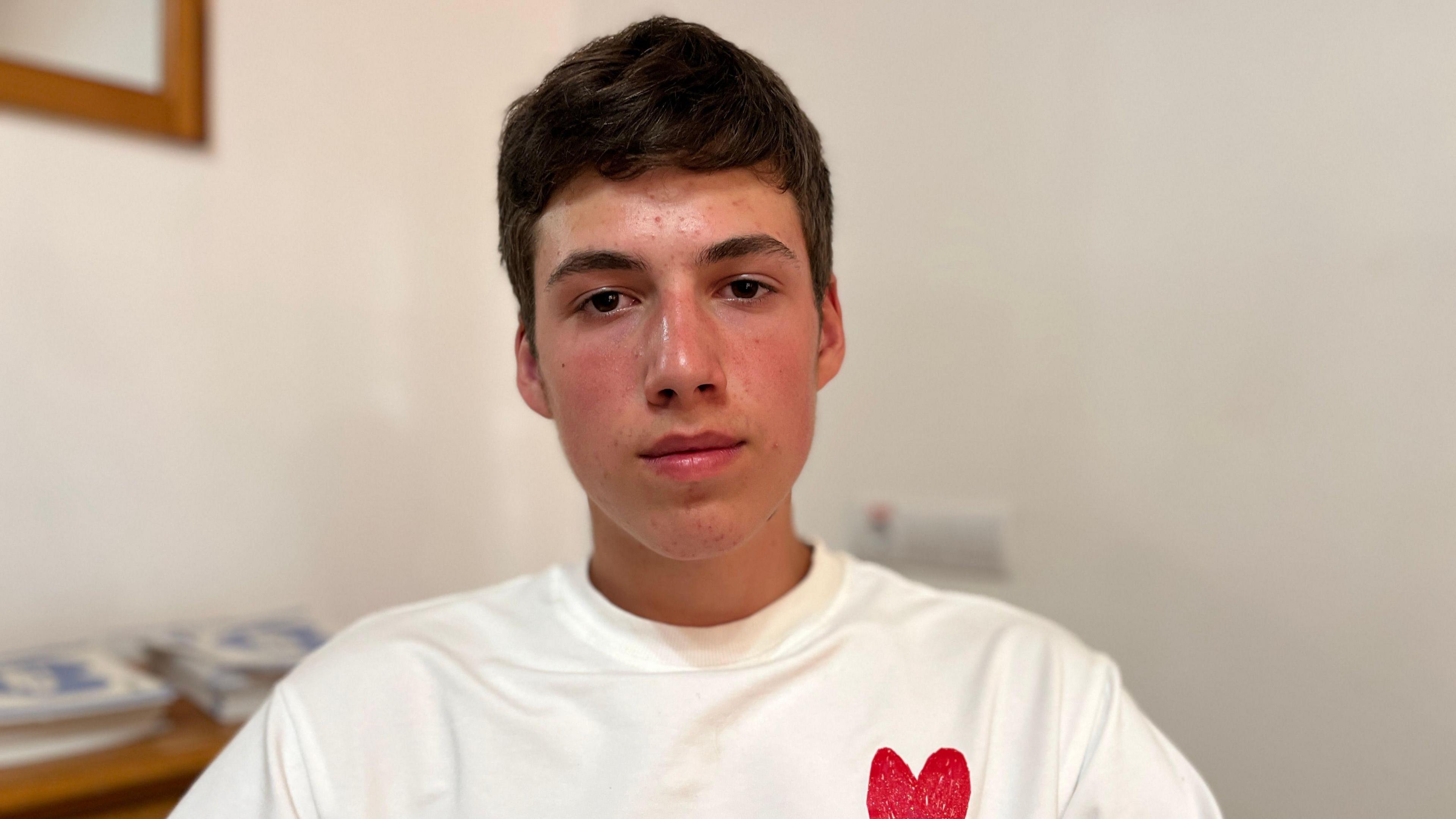 A teenage boy looks straight faced into the camera , posing for a picture, wearing a white t-shirt featuring a small red heart logo