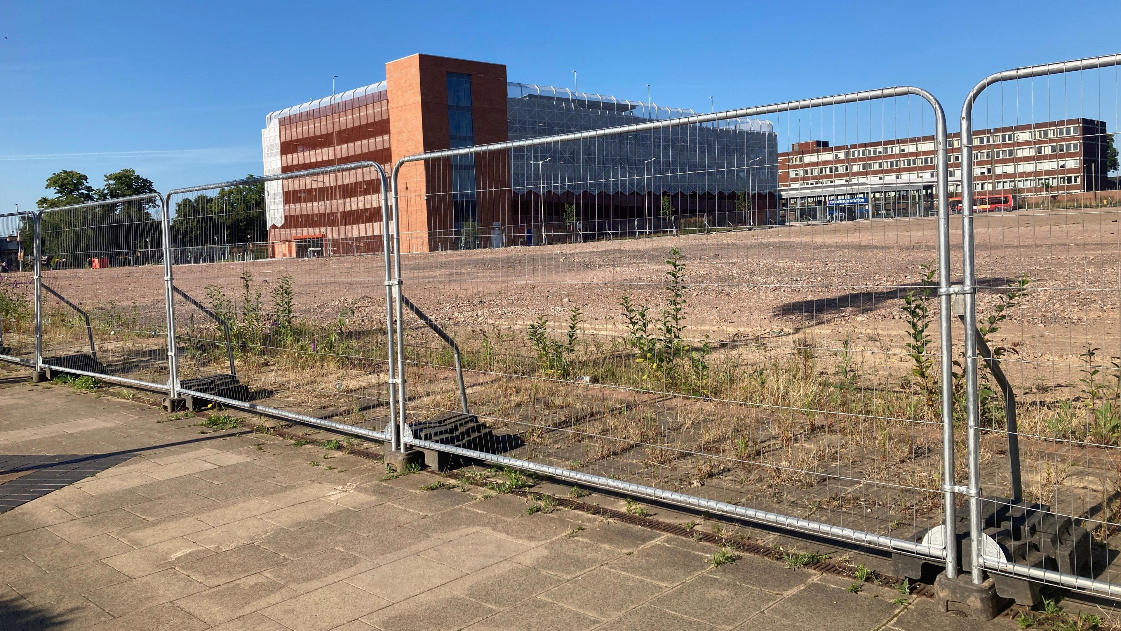 The empty Royal Arcade site in Crewe. There are metal temporary railings outside a bare patch of land with some green weeds growing behind them. In the distance is the brick and steel outside of a building behind the land.