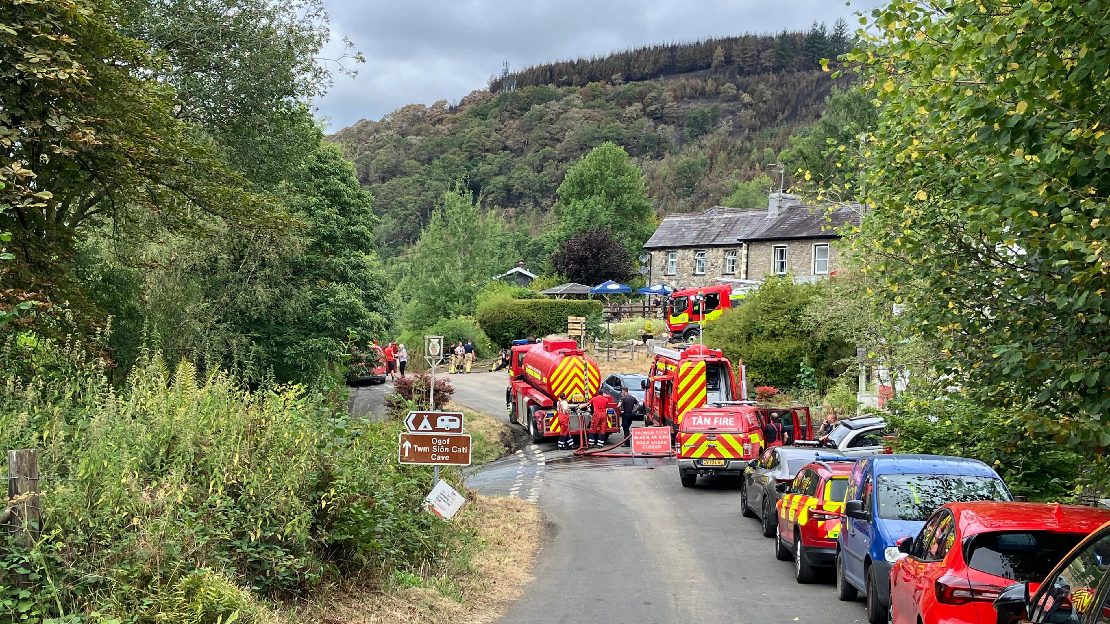Fire trucks can be seen at the bottom of a hill on a cordoned off road. There are fire crews stood around it.